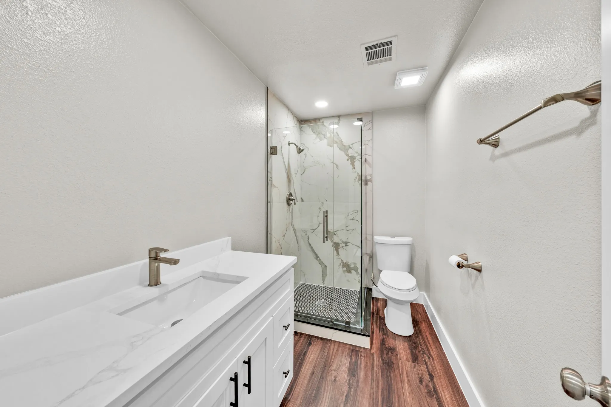 White shaker vanity with black hardware, white countertop, bronze-finish faucet, glass-enclosed shower with tiled surround, and wood-finish flooring