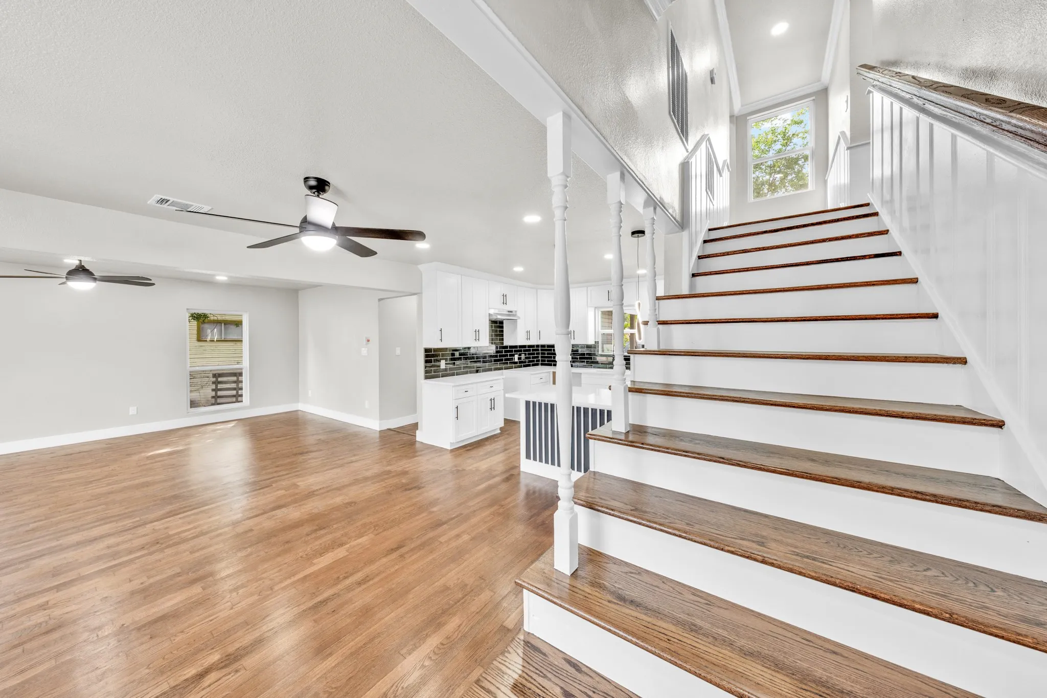 Wood-finish flooring throughout the main living area, a white cabinet kitchen with dark tile backsplash, a two-tone staircase with wood treads and white risers, and multiple ceiling fans