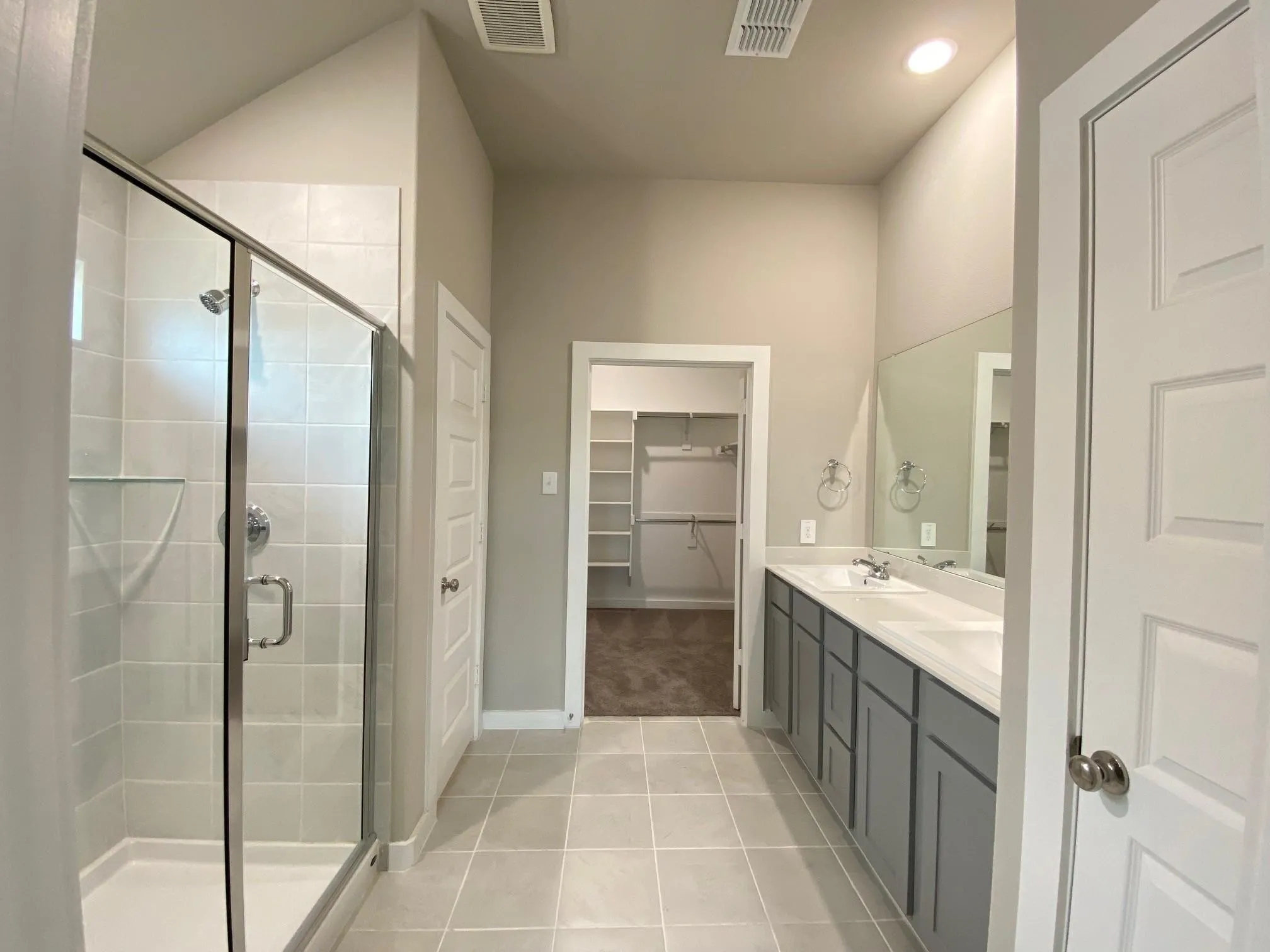 Walk-in shower with clear glass enclosure and tiled walls, dual vanity with undermount sinks, gray cabinetry, and a full-width mirror