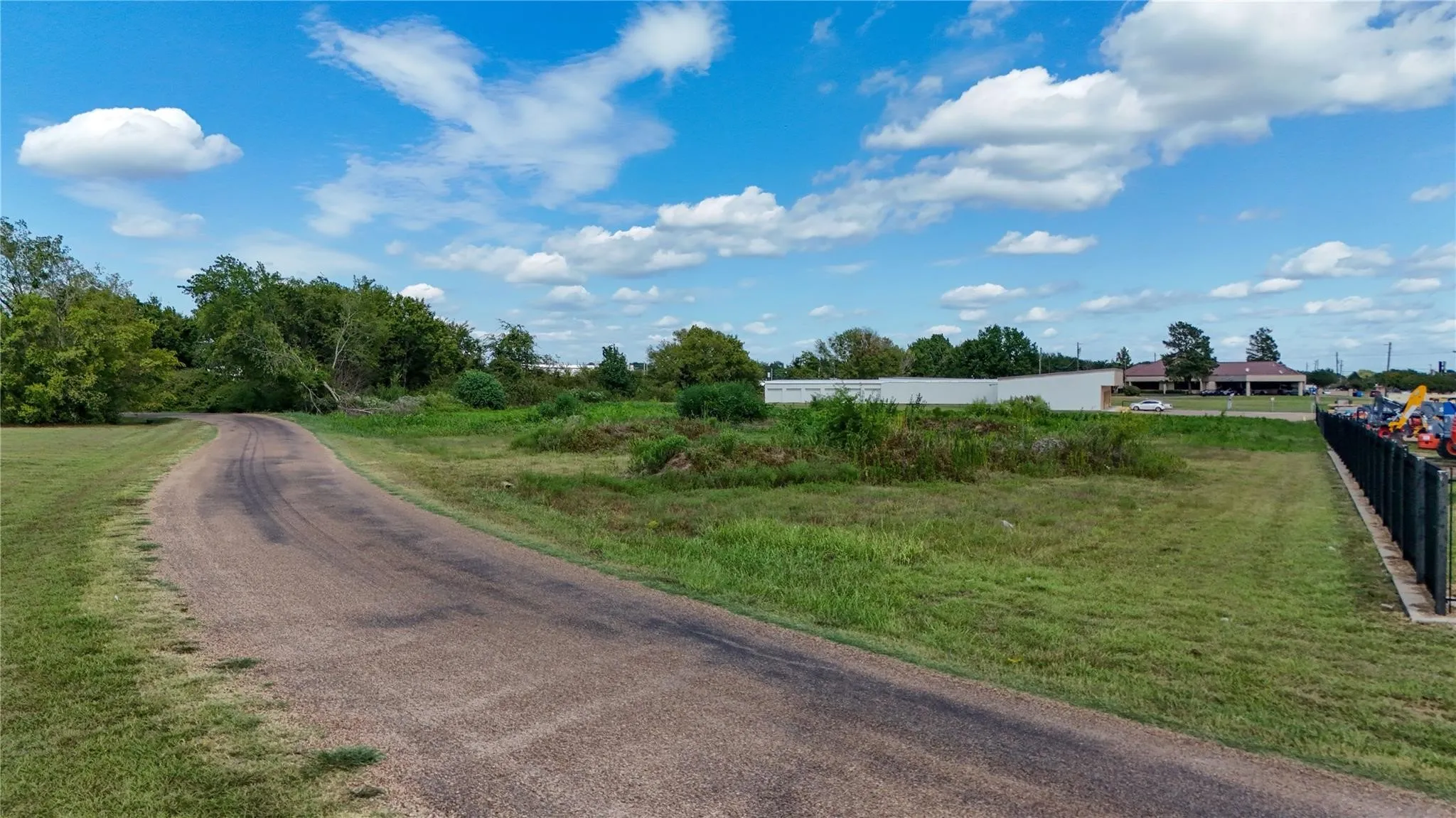 Paved access road, extensive green space, mature trees, security fence, and distant commercial buildings