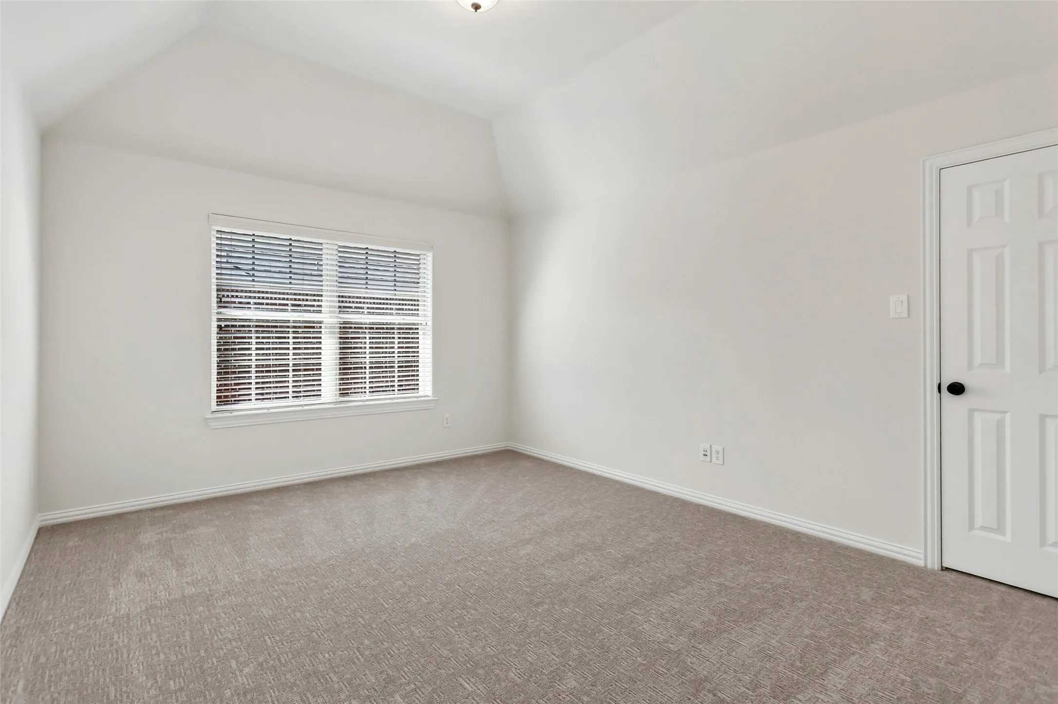 2nd floor bedroom. Vaulted ceiling, neutral tone walls, carpet flooring, window with horizontal blinds, and a paneled door with black hardware