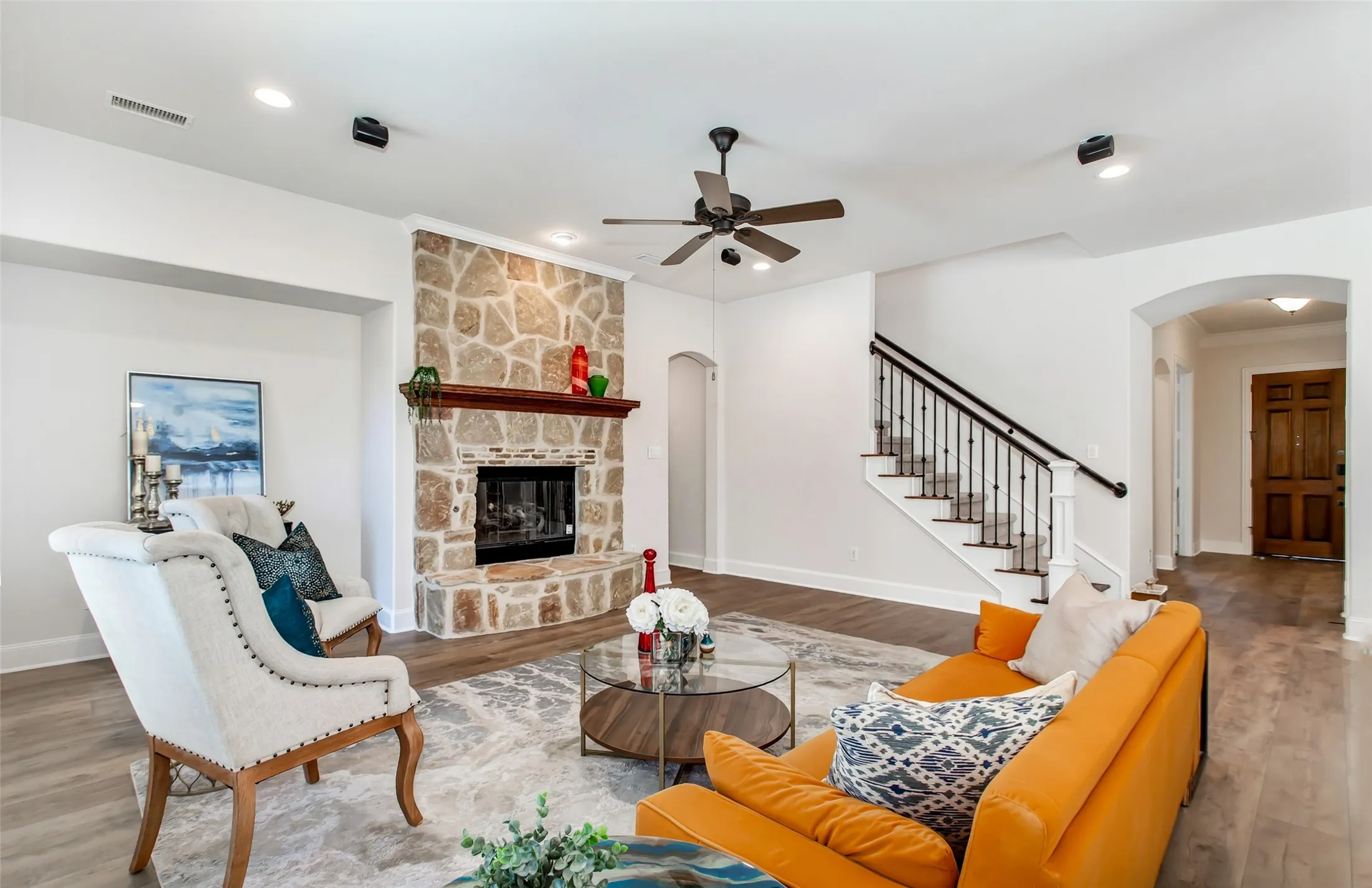Stone fireplace with a wood mantel, wood-finish flooring, staircase with wrought iron balusters, arched entryway, and a ceiling fan