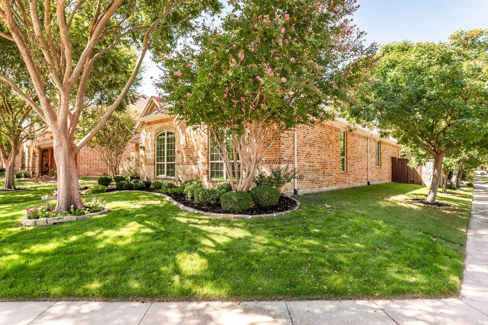 Brick and stone exterior with arched windows, manicured landscaping, mature trees, and a concrete sidewalk