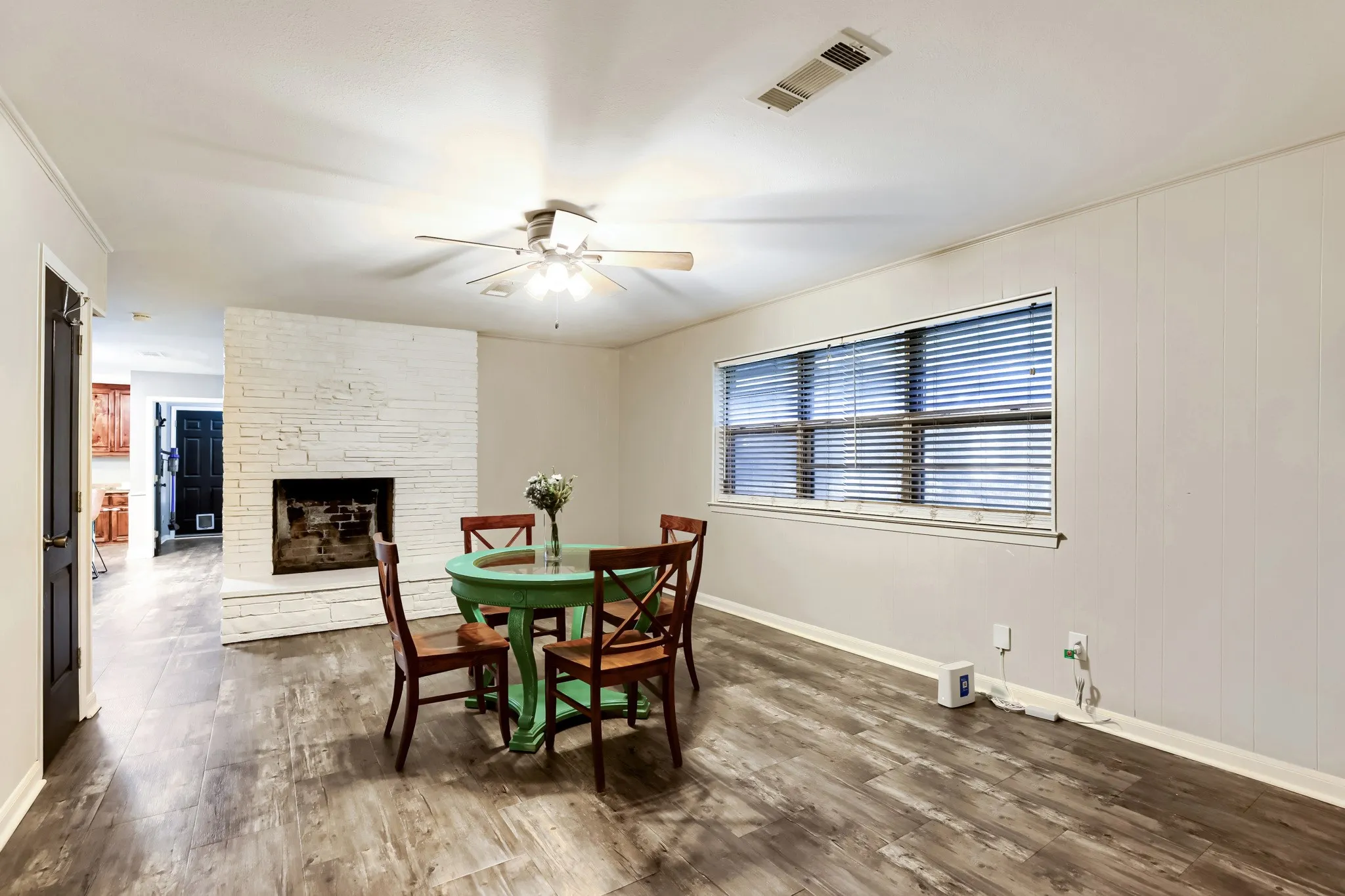 Wood-finish flooring, white brick fireplace, ceiling fan, and a large window with blinds