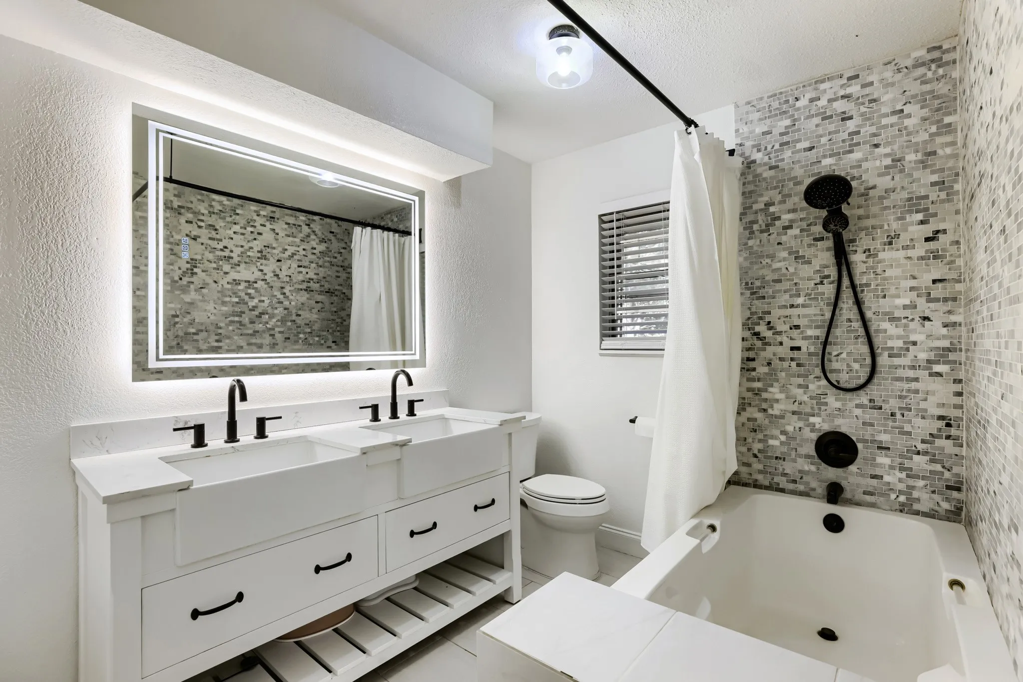 Dual vanity with integrated sinks, back-lit mirror, matte black fixtures, and mosaic tile shower surround
