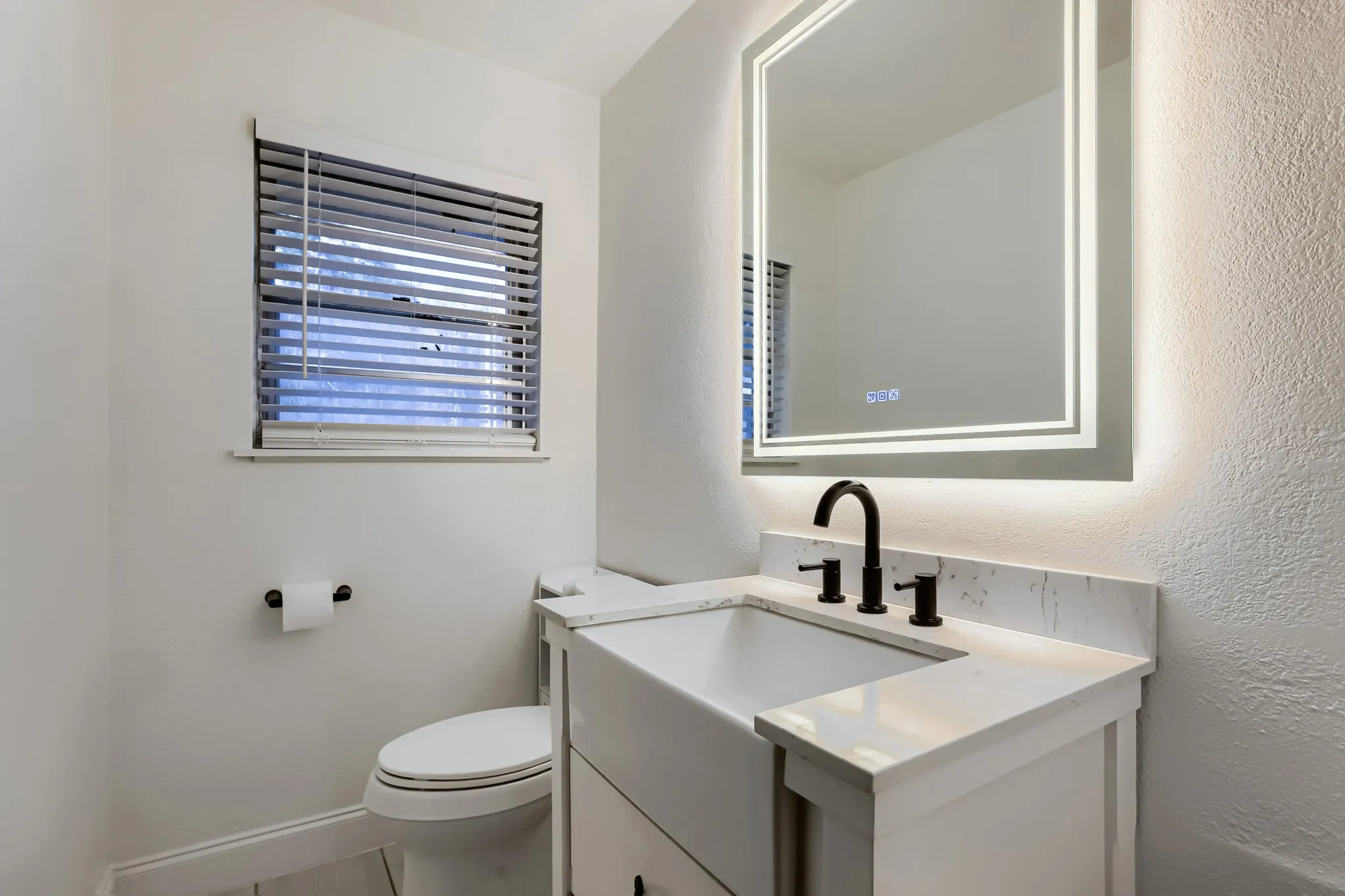 White vanity with an integrated farmhouse-style sink, solid surface countertop, matte black faucet, and an illuminated mirror