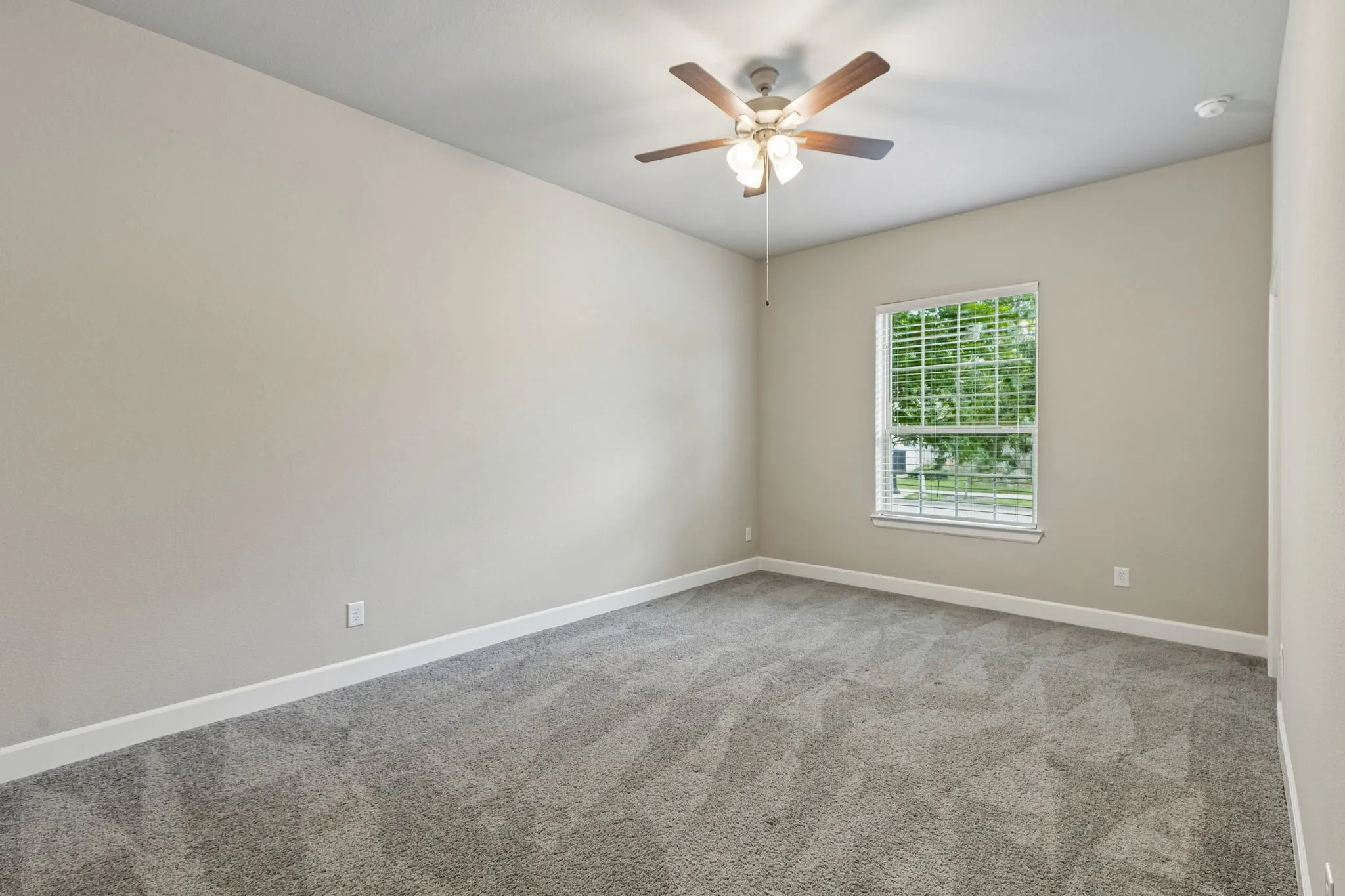 Bright corner bedroom featuring neutral wall finishes, plush carpet flooring, a ceiling fan with integrated lighting, and natural light from a large double-hung window with plantation shutters
