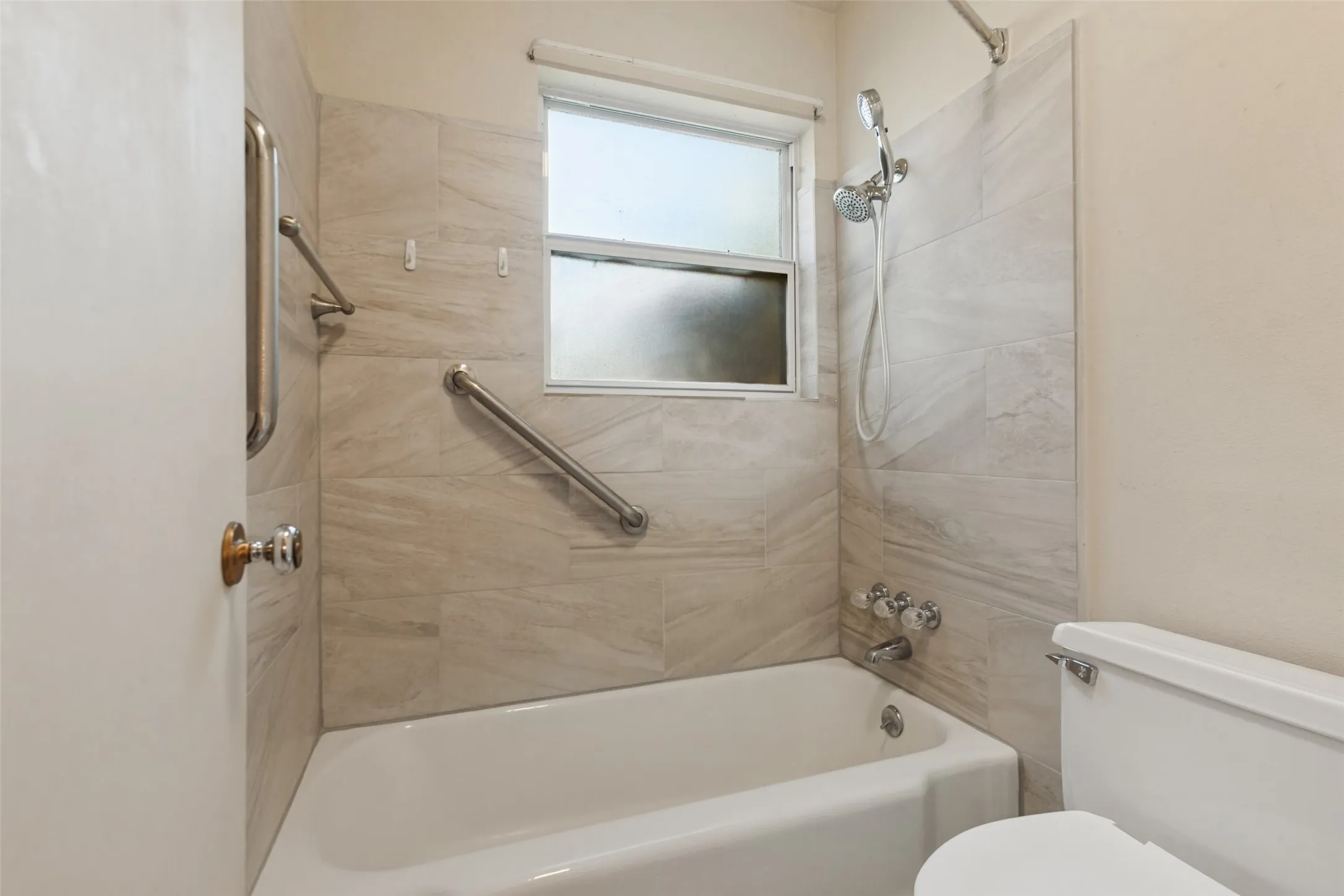 Bathtub with a shower fixture, wall-mounted grab bars, tiled shower surround, and a window for natural light