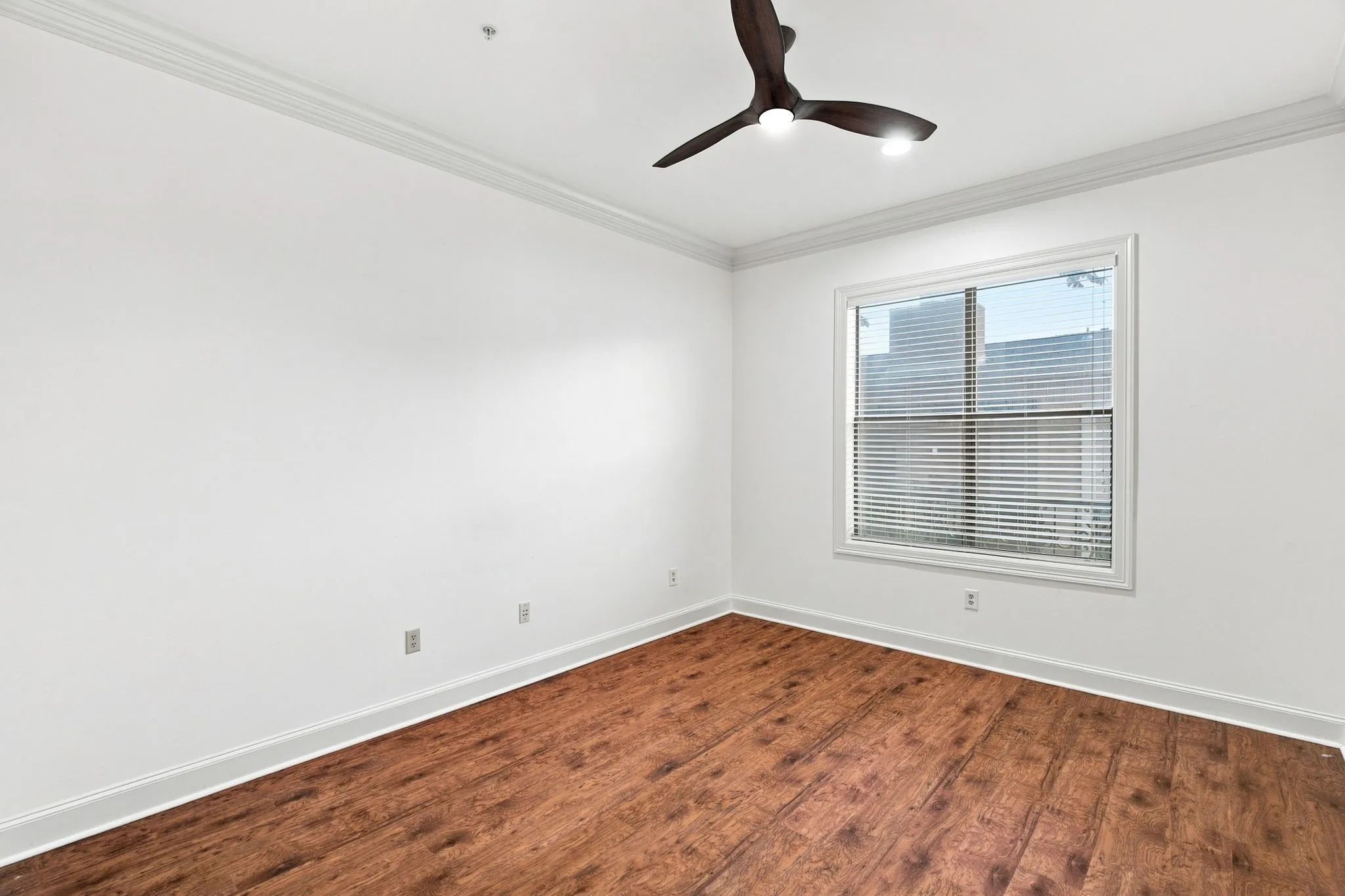 Guest bedroom with wood-finish flooring, white walls, crown molding, a window with blinds, and a dark-toned ceiling fan with integrated lighting
