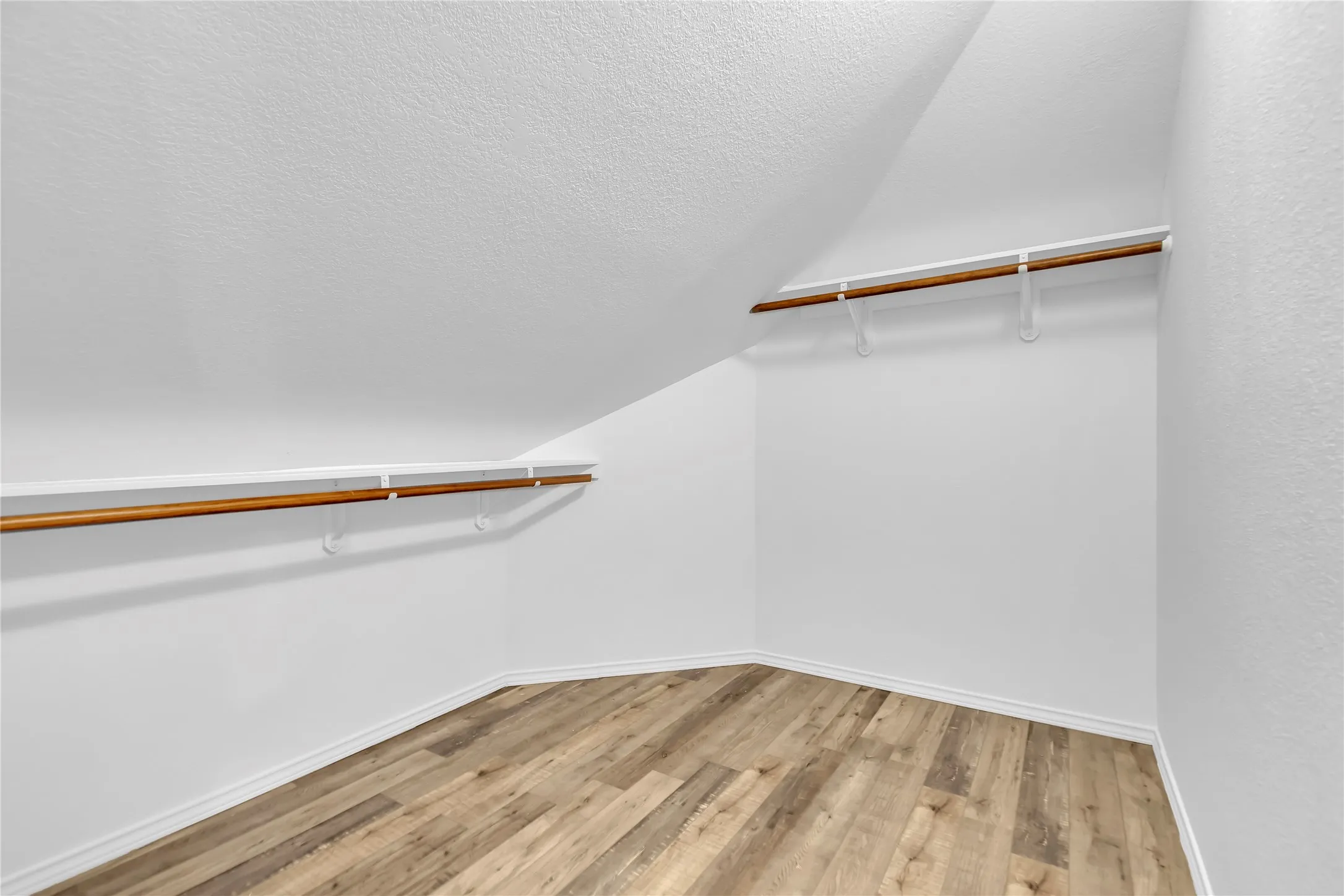 Angled ceiling architecture, wood-finish flooring, white baseboards, and built-in shelving with hanging rods