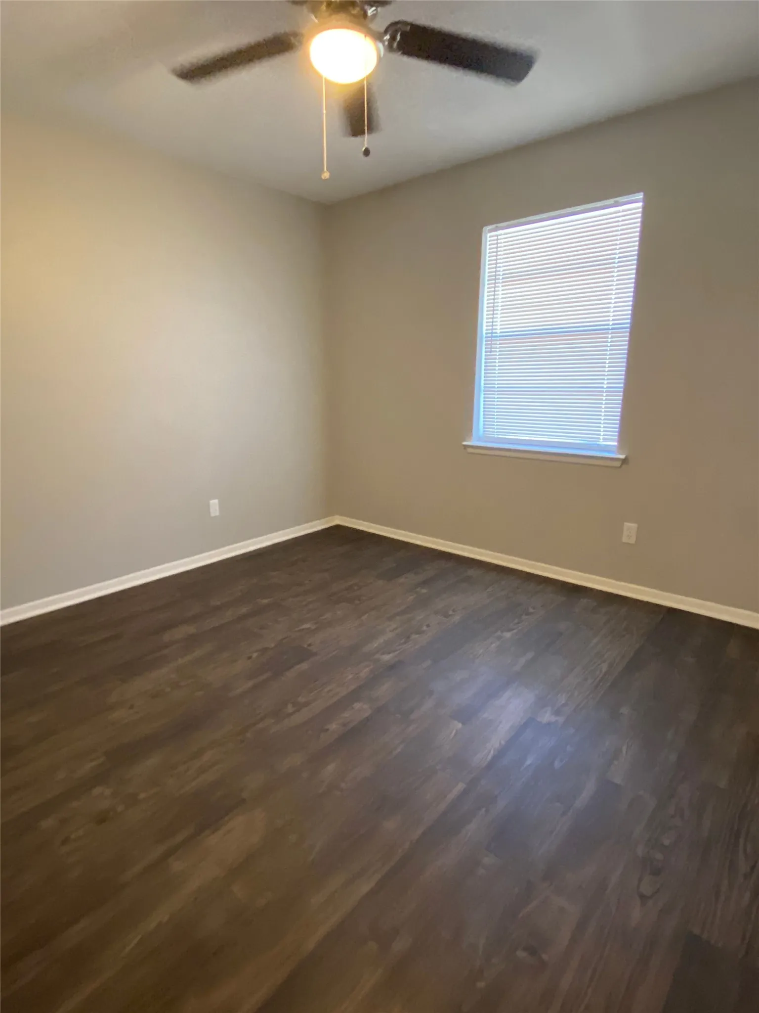 Spacious room featuring wood-finish flooring, a ceiling fan with integrated lighting, and a window with blinds