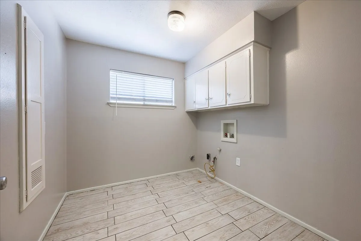 White cabinetry with clear knobs, window with blinds, wood-finish tile flooring, ceiling-mounted light fixture, and built-in utility connections