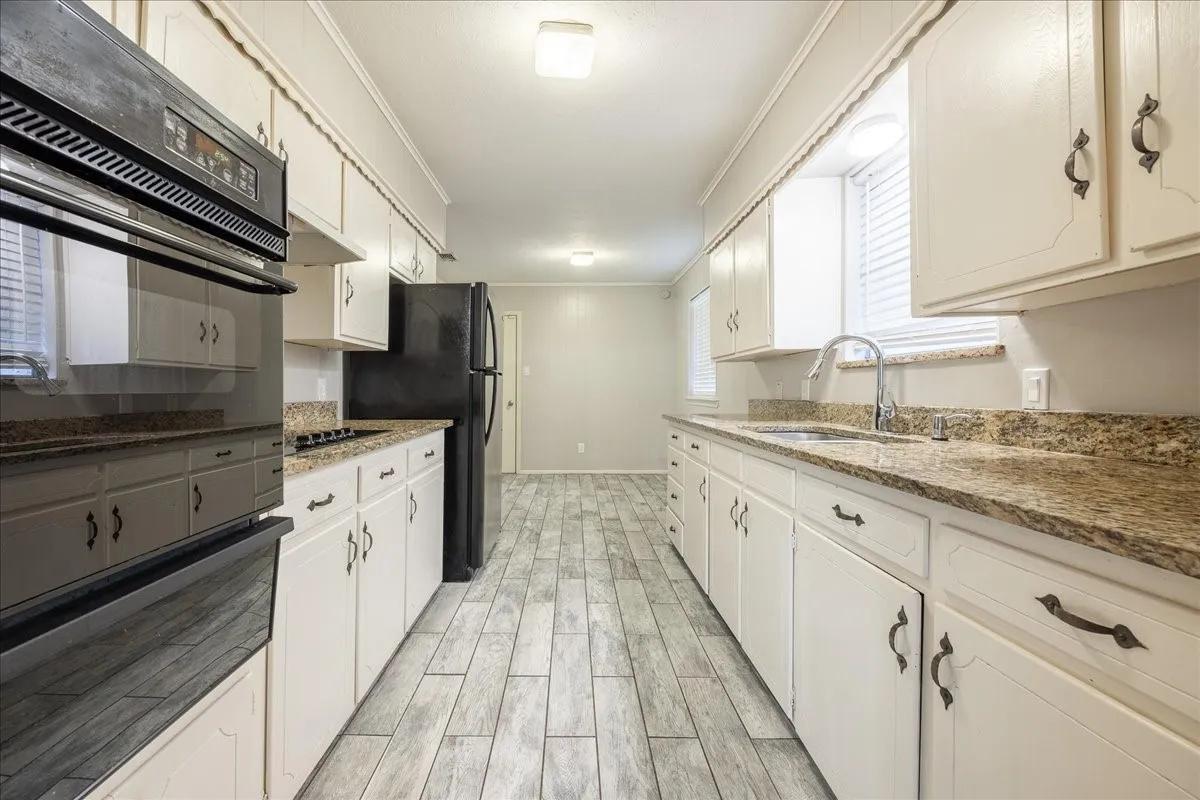 Galley kitchen featuring wood-finish flooring, white cabinetry with dark hardware, stone-finish countertops, a double-basin sink with a gooseneck faucet, and integrated appliances