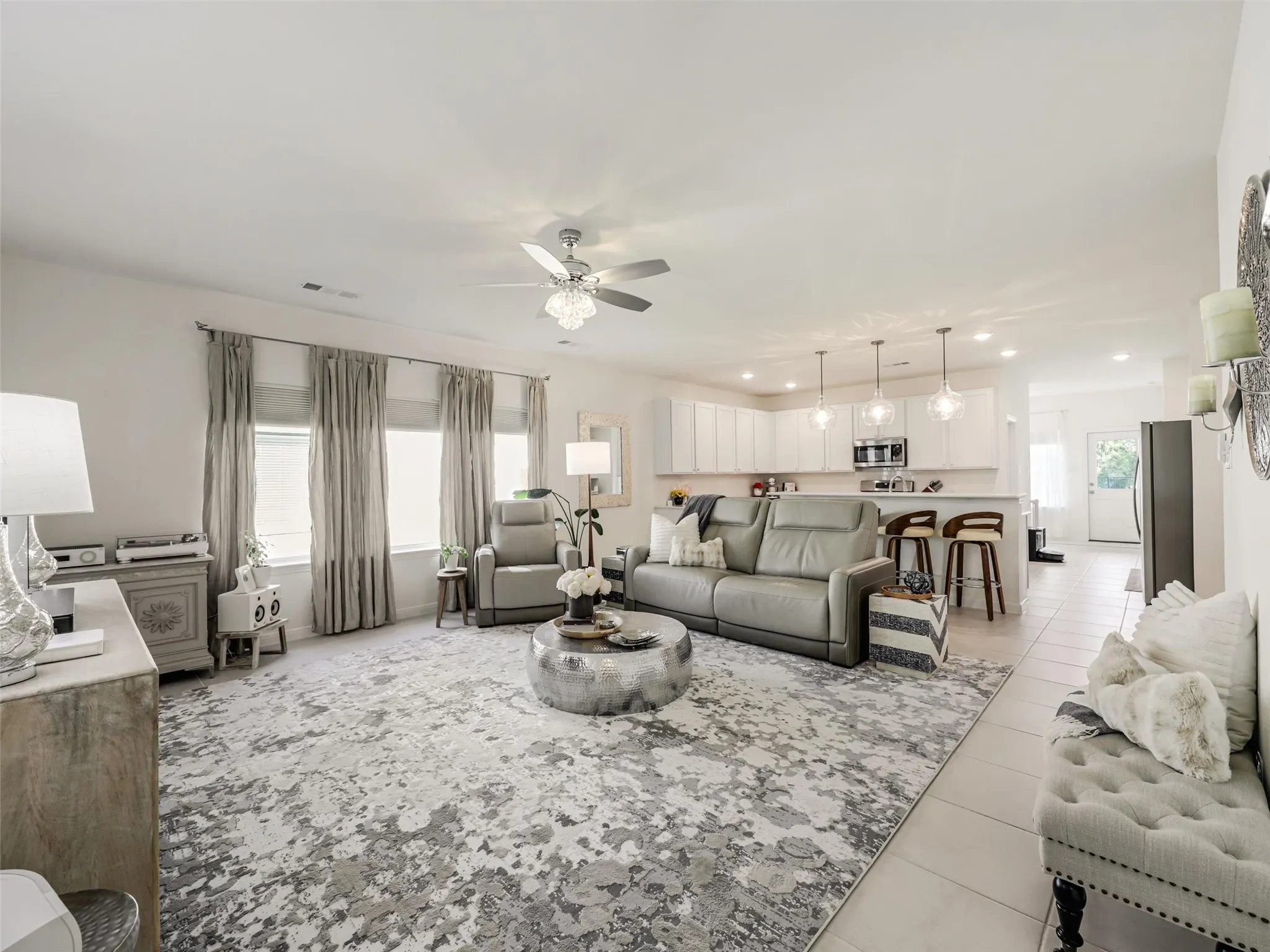 Open-concept living area featuring a ceiling fan with light fixture, three large windows, a breakfast bar with pendant lighting, and ceramic tile flooring
