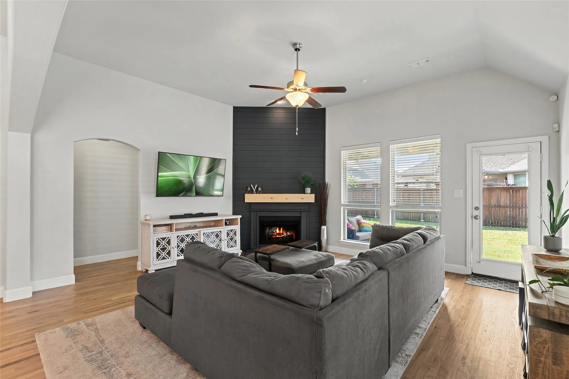 Vaulted ceiling with a ceiling fan, fireplace with a wood mantle and dark shiplap surround, wood-finish flooring, multiple windows, and an arched doorway