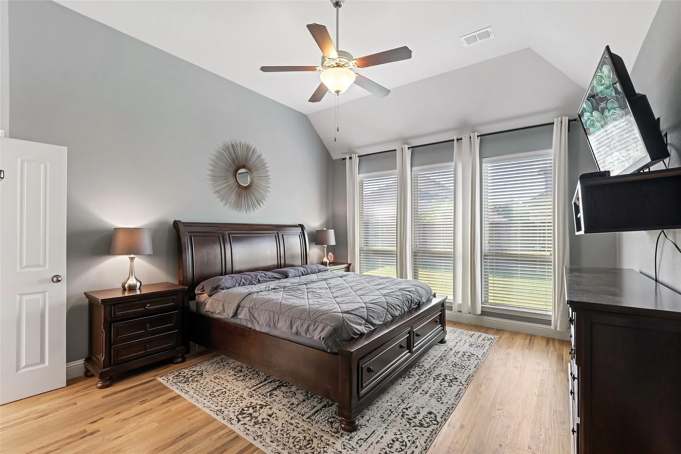 Vaulted ceiling with a ceiling fan, three large windows with blinds, light wood-finish flooring, and a built-in wall-mounted television bracket