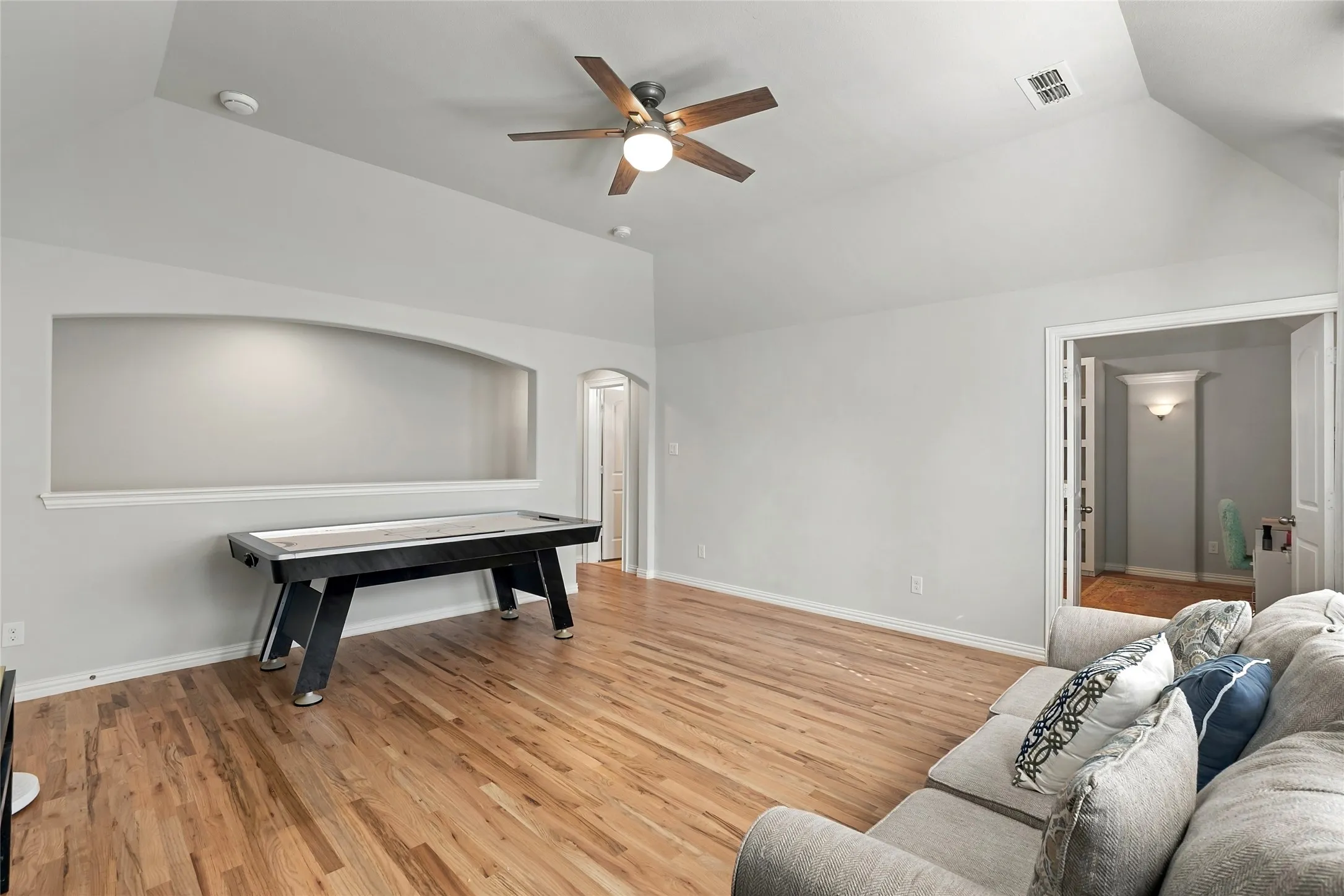 Vaulted ceiling with an integrated ceiling fan, arched wall niche with accent lighting, wood-finish flooring, and a cased doorway leading to an interior space