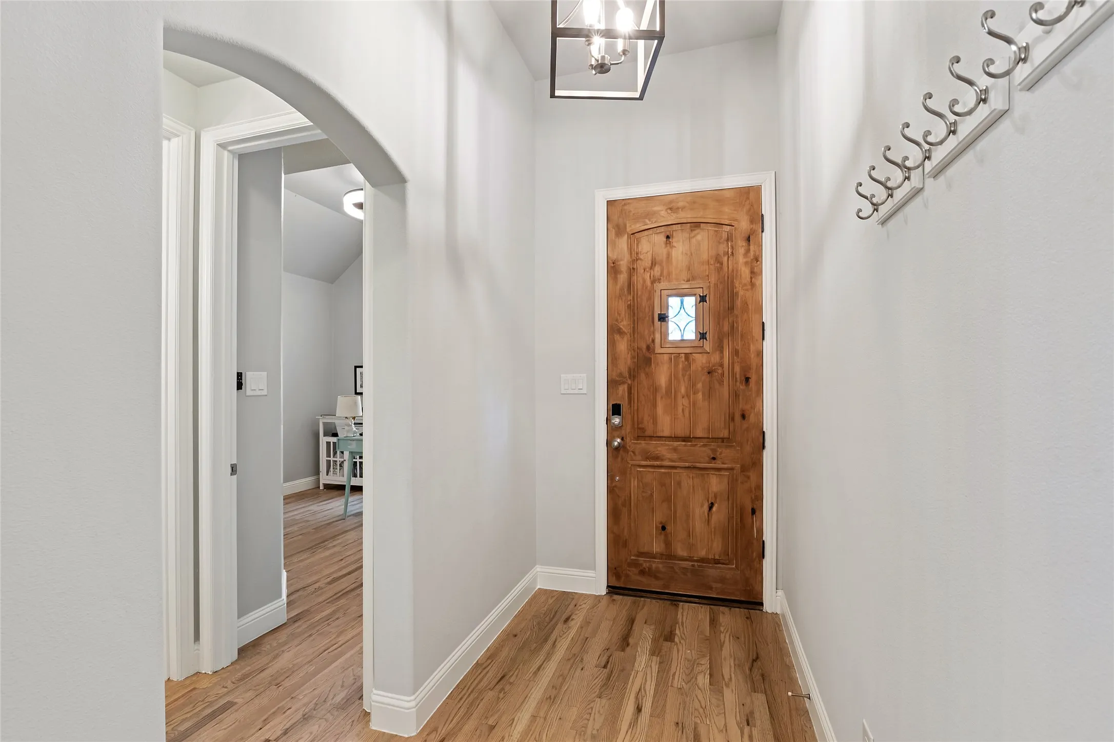Wood-finish flooring throughout, solid wood entry door with decorative glass insert, ceiling-mounted light fixture, and archway entry to an additional room