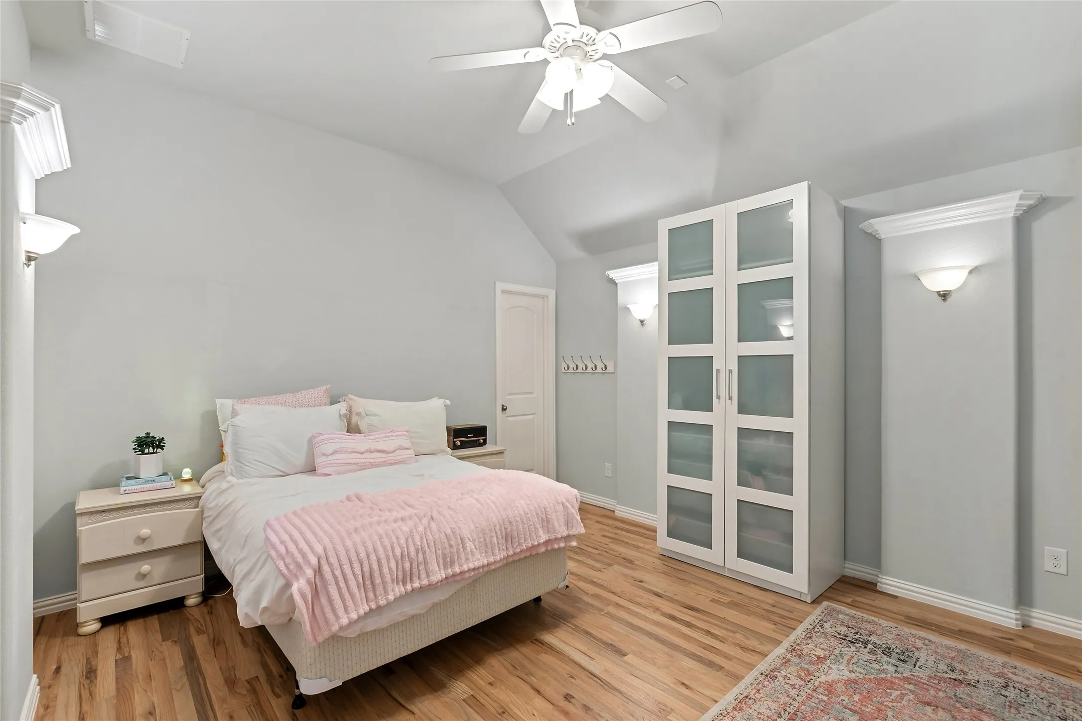 Vaulted ceiling with a ceiling fan, recessed wall sconces with crown molding, wood-finish flooring, and a white storage cabinet with frosted glass doors