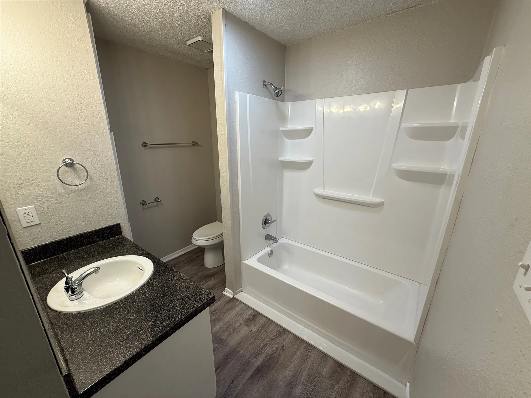 Bathroom featuring a white tub, integrated shower surround with shelving, dark countertop with an integrated sink, and wood-finish flooring