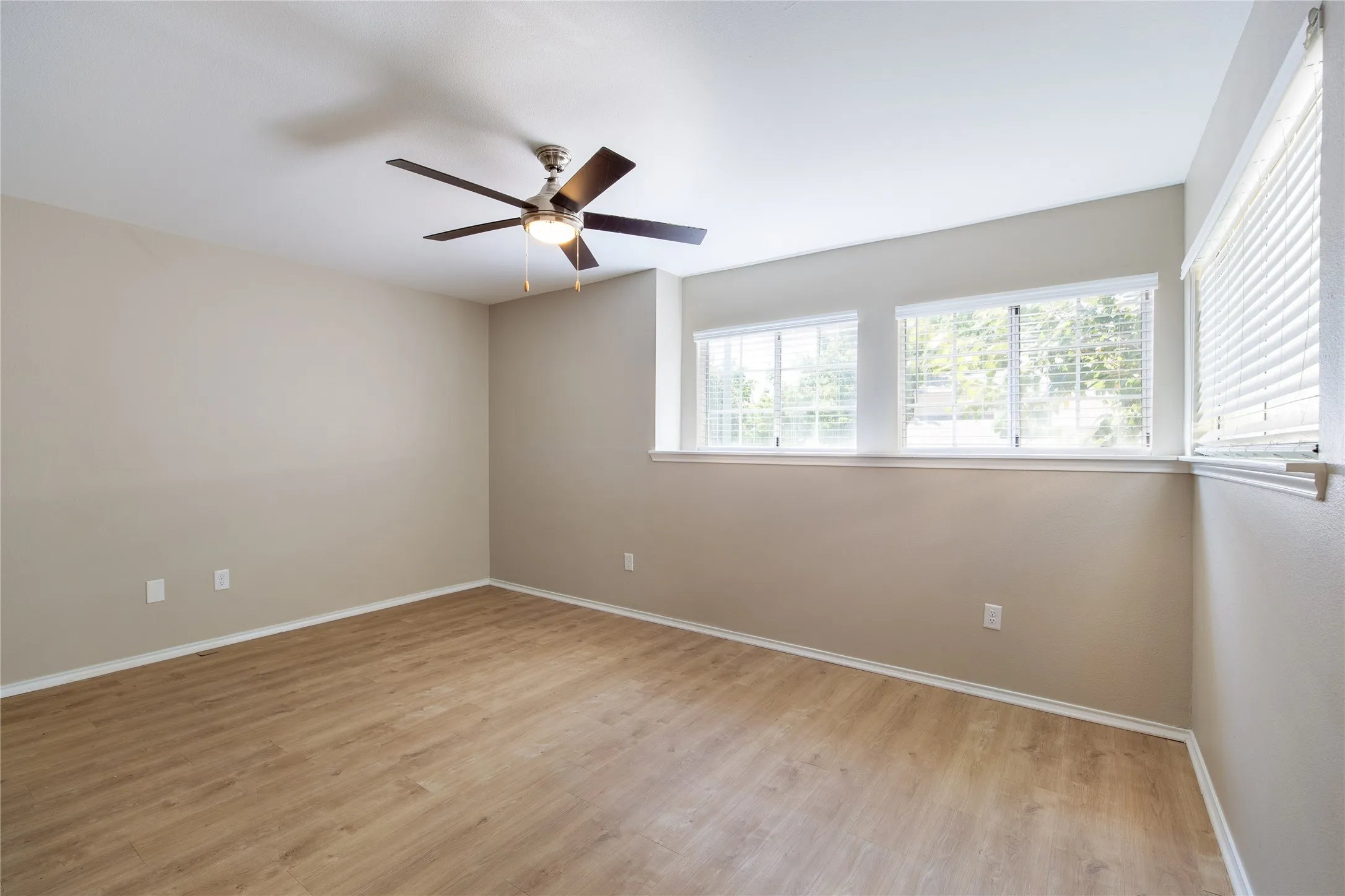 Wood-finish flooring, neutral wall tones, ceiling fan with integrated lighting, and multiple windows with blinds