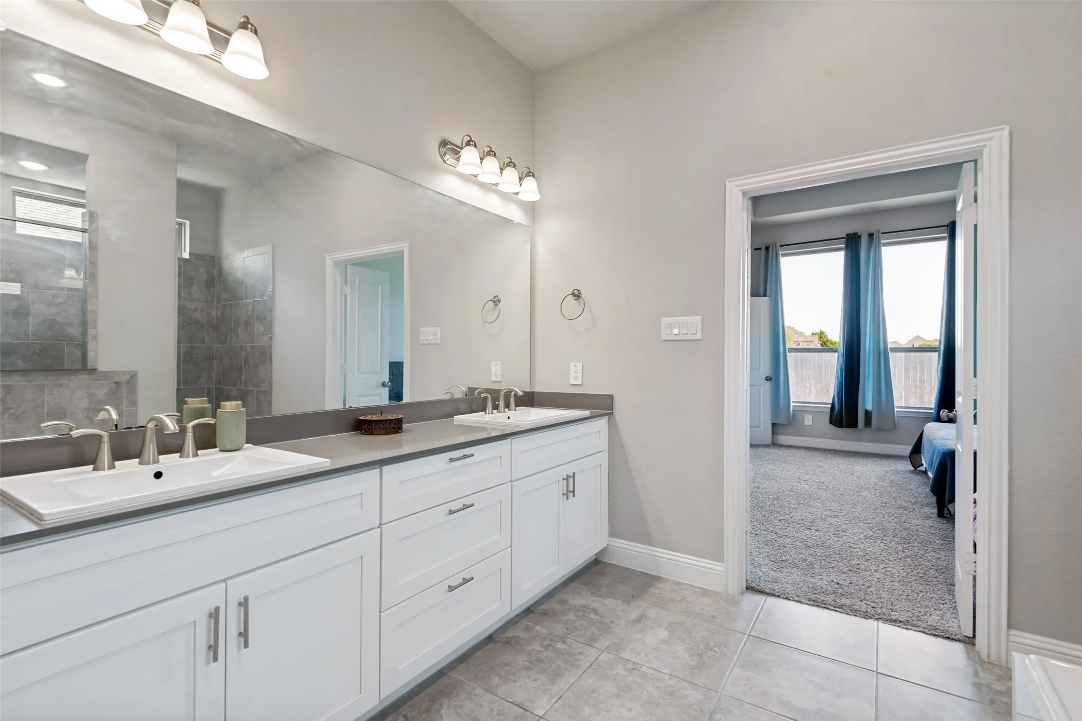 Bathroom featuring dual rectangular undermount sinks, white shaker-style vanity cabinetry, a long wall-mounted mirror, and tile flooring