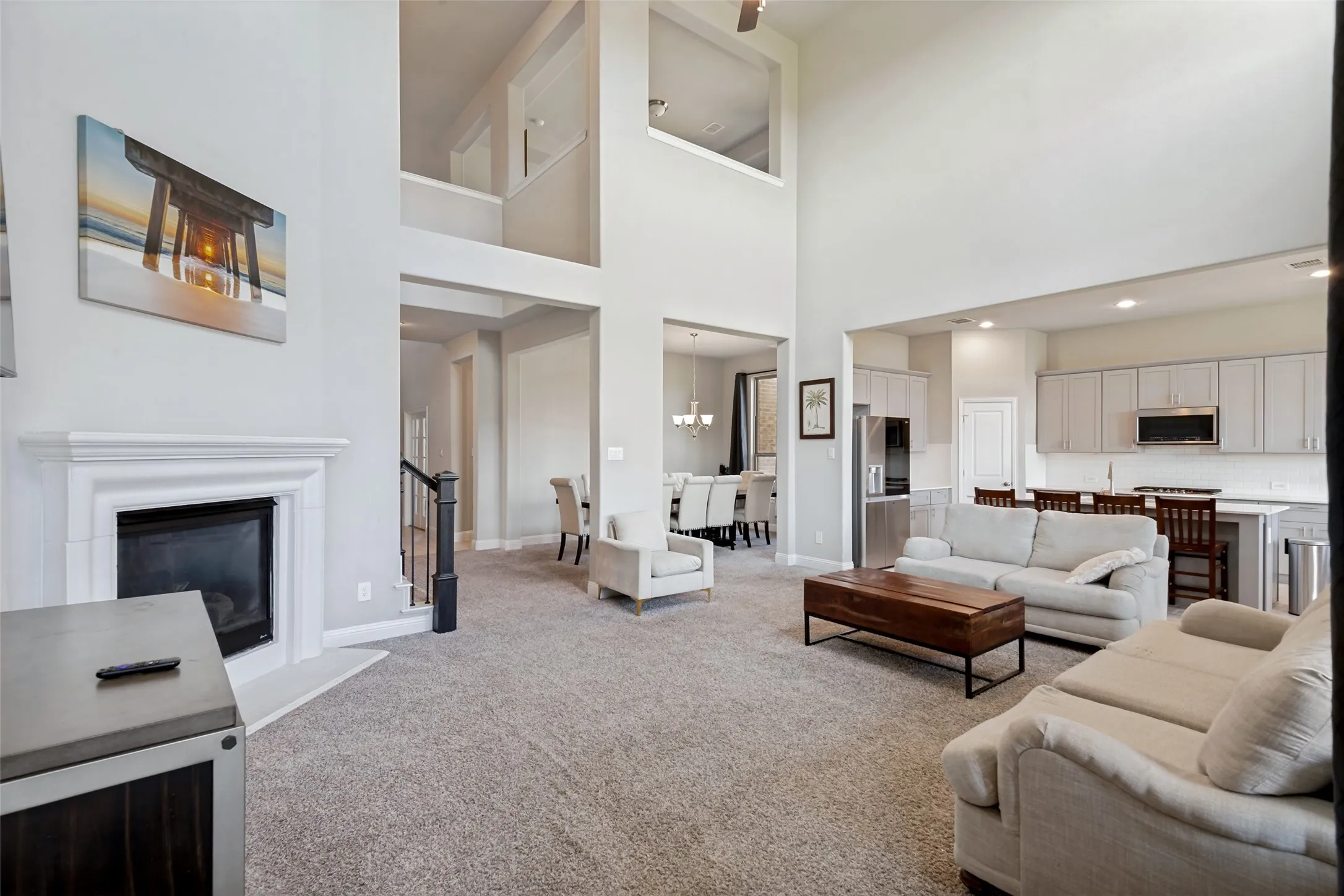Two-story ceiling, integrated kitchen and living area, fireplace with hearth, recessed lighting, and a staircase with a dark wood-finish handrail
