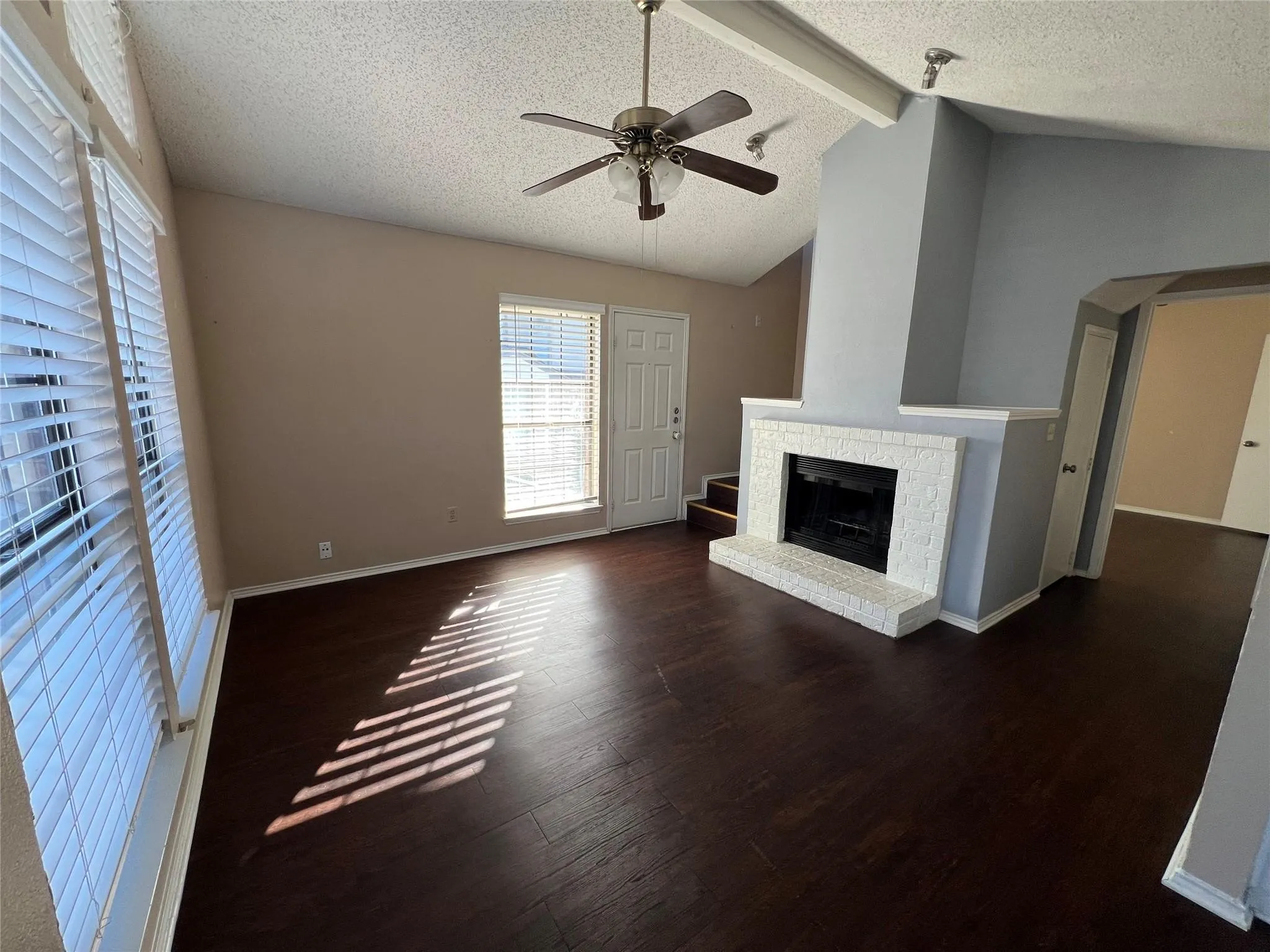 Wood-finish flooring, vaulted ceiling, white brick fireplace with hearth, and ceiling fan with integrated lighting