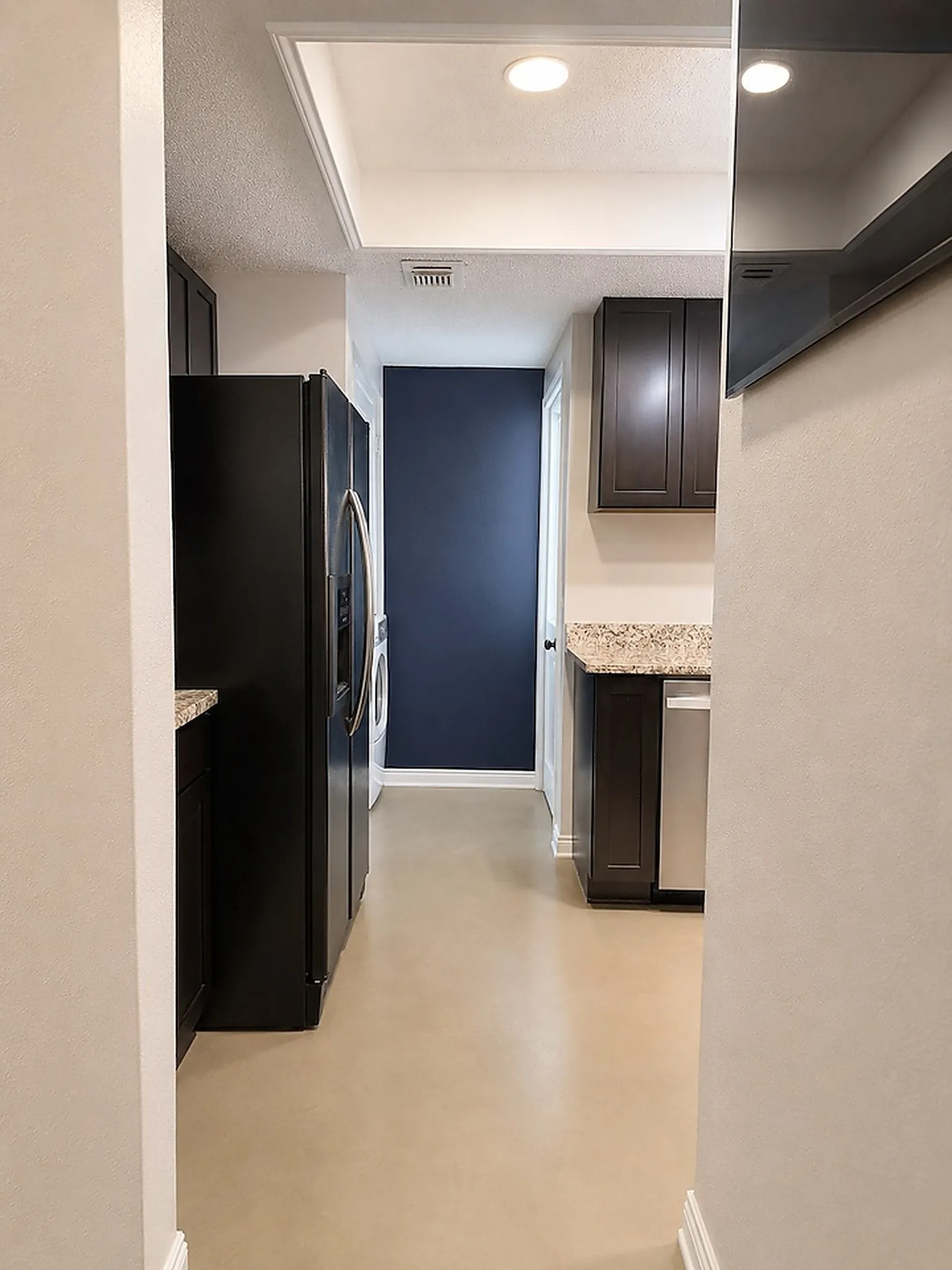 Recessed lighting illuminates the galley-style kitchen featuring dark wood-finish cabinetry, light-toned countertops, and a stainless steel dishwasher