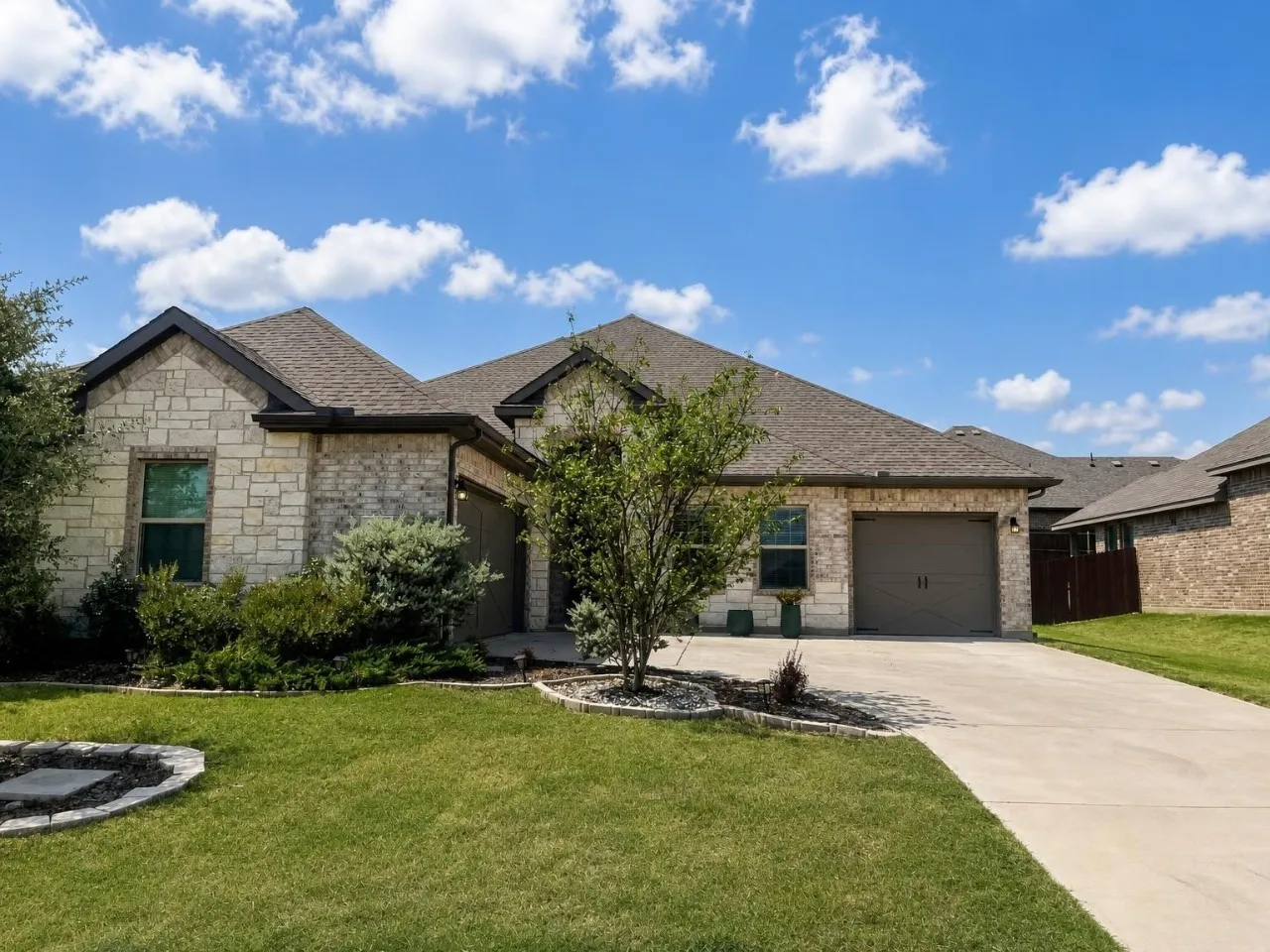 Stone and brick-veneer exterior with a shingled roof, attached garage, concrete driveway, and established front landscaping