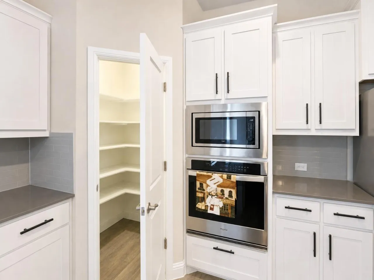 White shaker-style cabinetry with matte black hardware, stainless steel built-in microwave and oven, gray subway tile backsplash, and a walk-in pantry with wood-finish flooring