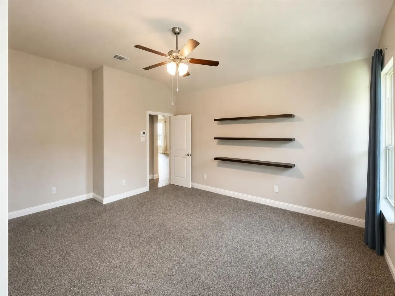 Spacious room featuring neutral carpeting, a ceiling fan with integrated lighting, and three wall-mounted floating shelves