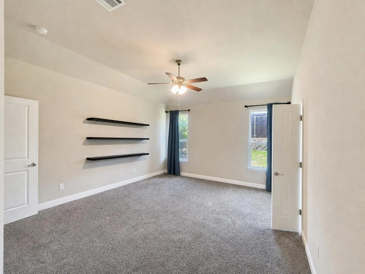 Vaulted ceiling with ceiling fan, two windows with curtain rods, three floating shelves, light gray carpet, and white interior doors