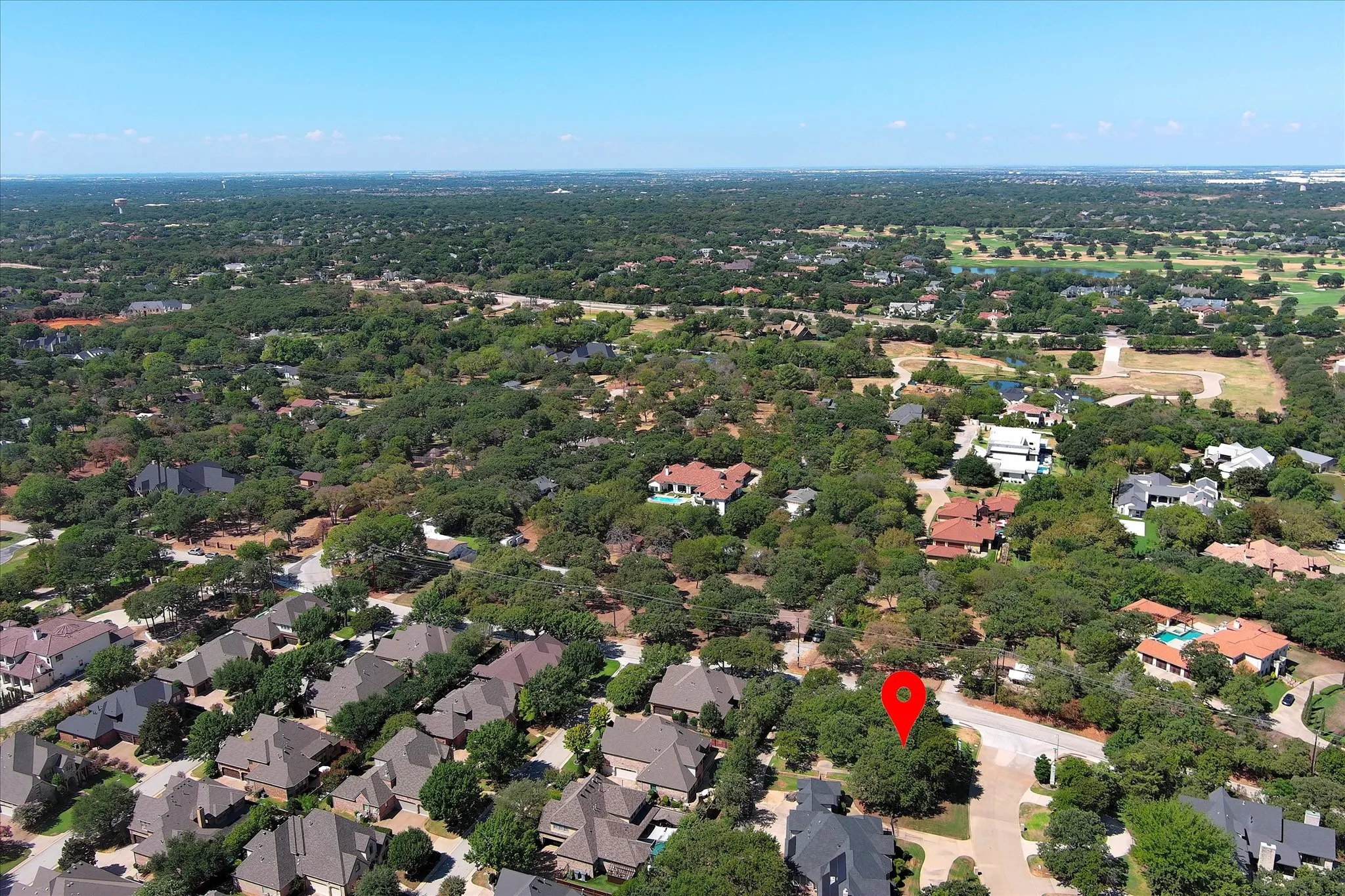 Aerial view shows heavily forested another direction shows Red marker on property location with upscale townhome community across the way and SH 114 in the distance above near top of photo.