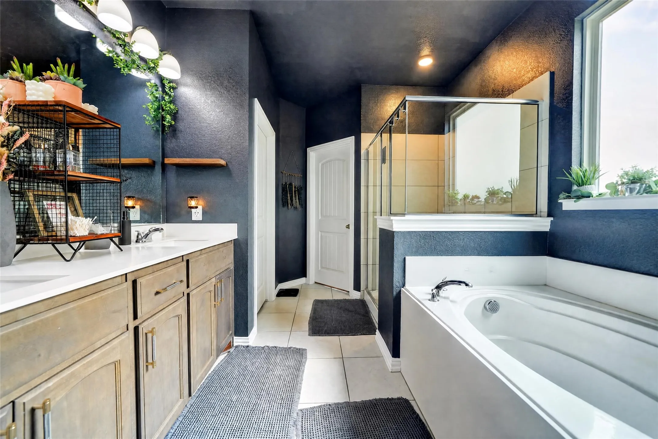 Dual vanity with white countertops, built-in soaking tub, glass-enclosed shower, and white tile flooring