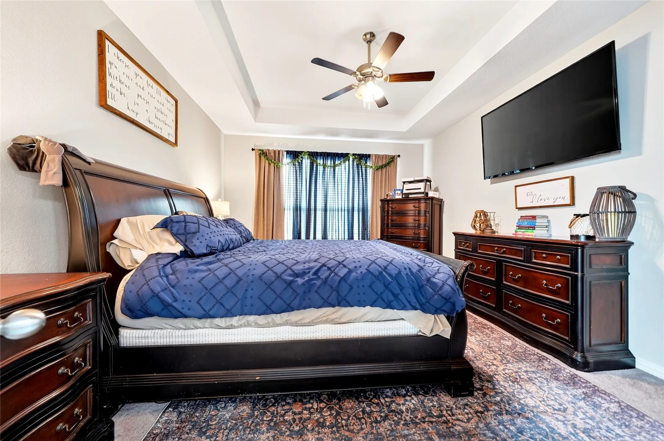 Primary Bedroom featuring a wood-frame sleigh bed, layered bedding in navy blue tones, dual wood nightstands with table lamps, a wood dresser with multiple drawers, ceiling fan with brushed metal accents, wall-mounted flat-screen television, recessed lighting, and patterned area rug anchoring the space