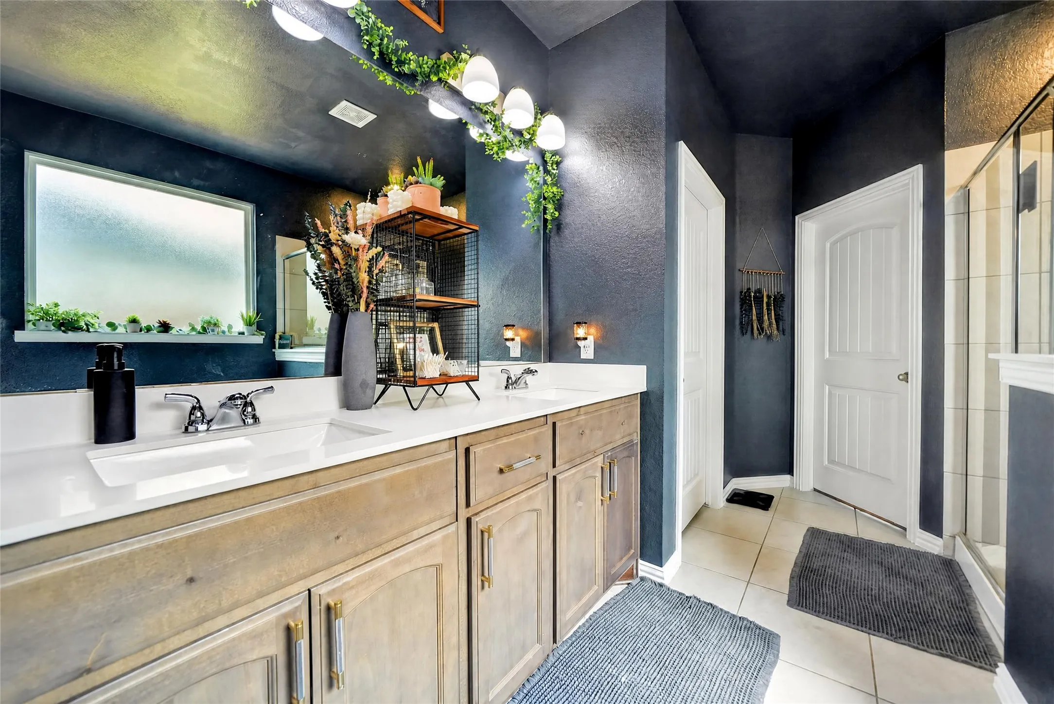 Dual sinks with chrome fixtures, wood-finish cabinetry, white countertops, textured blue walls, and a glass-enclosed shower