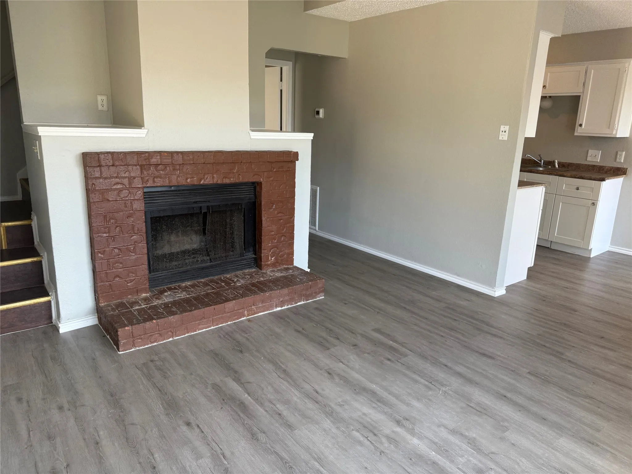 Fireplace with brick surround, wood-finish flooring, and white kitchen cabinetry