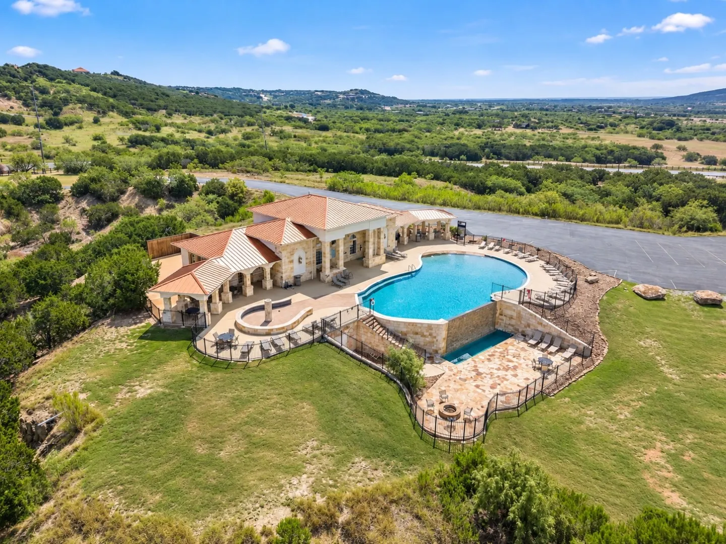 Resort-style pool with integrated spa and waterfall feature, expansive stone patio with built-in fire pit, covered outdoor lounging areas, and mature landscaping