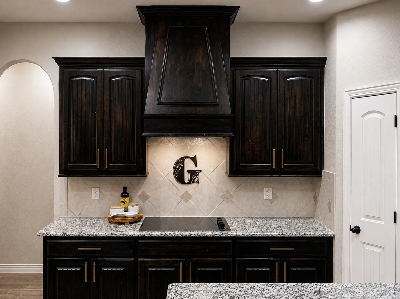 Dark wood cabinetry with crown molding, speckled countertops, tile backsplash, built-in cooktop, and an integrated range hood