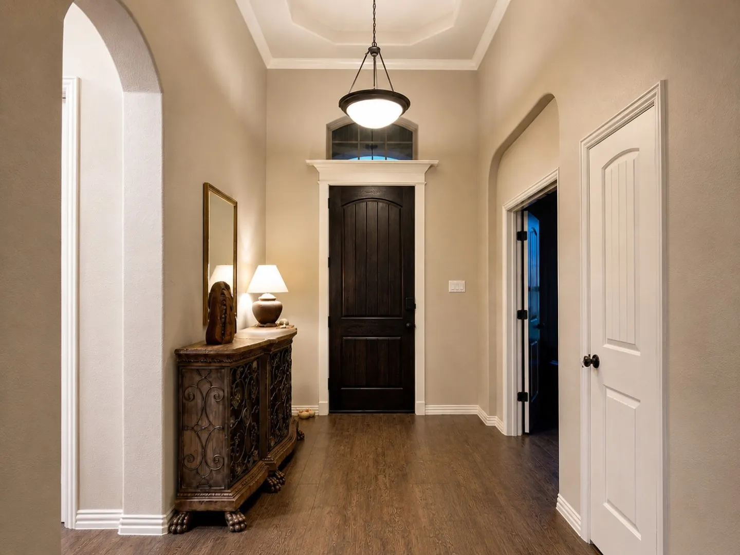 Wood-finish flooring throughout, tray ceiling with crown molding, dark wood front door with transom window, and arched doorways