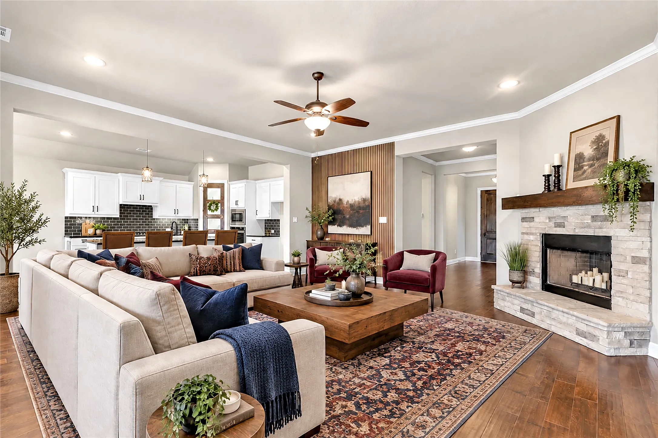 Stone-clad fireplace with a wood mantle, recessed lighting, ceiling fan, wide plank wood-finish flooring, and white crown molding. Virtual staged.