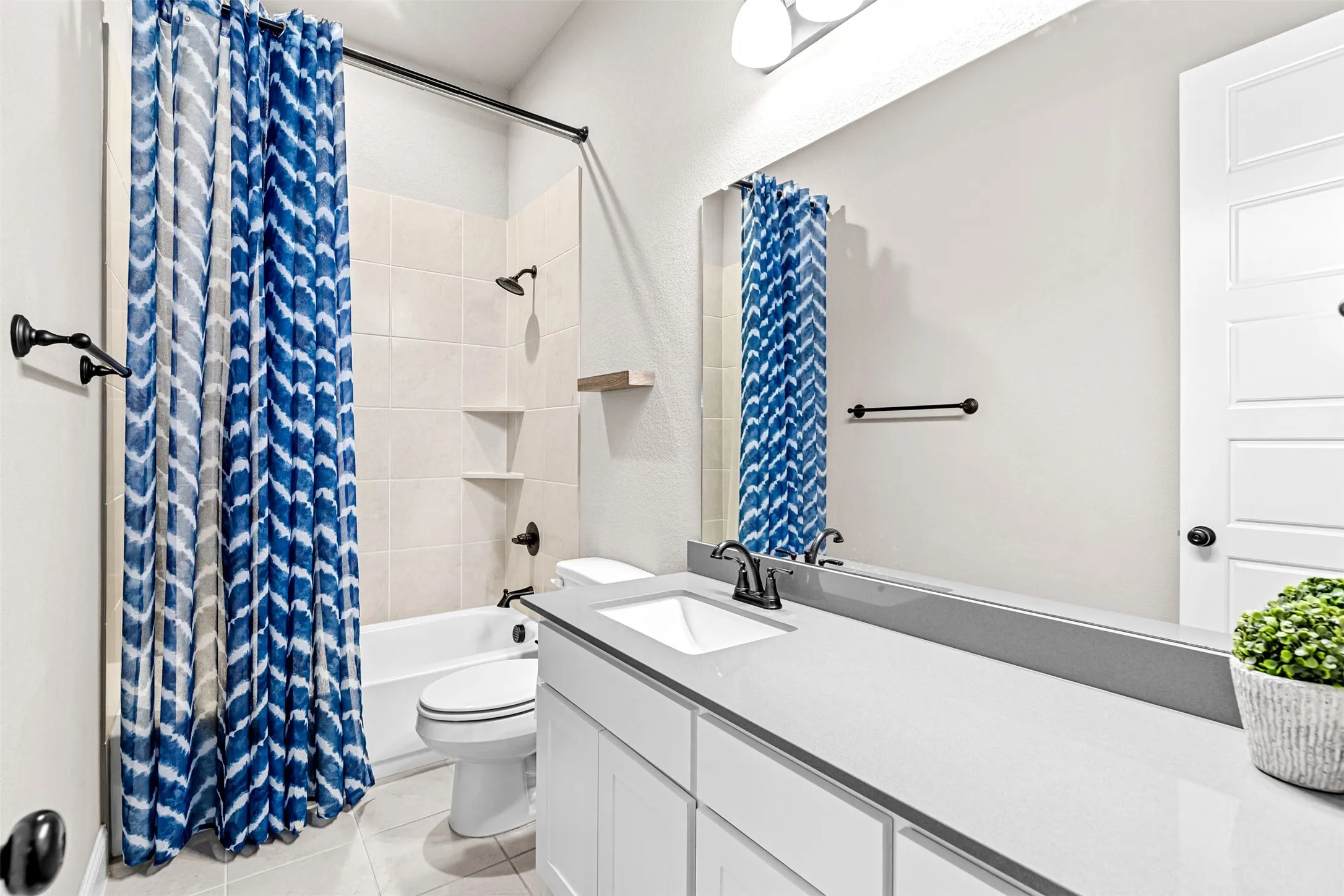 Full-width vanity mirror, white vanity cabinetry with dark hardware, white under-mount sink, dark faucet, and tile-surround shower with built-in shelving