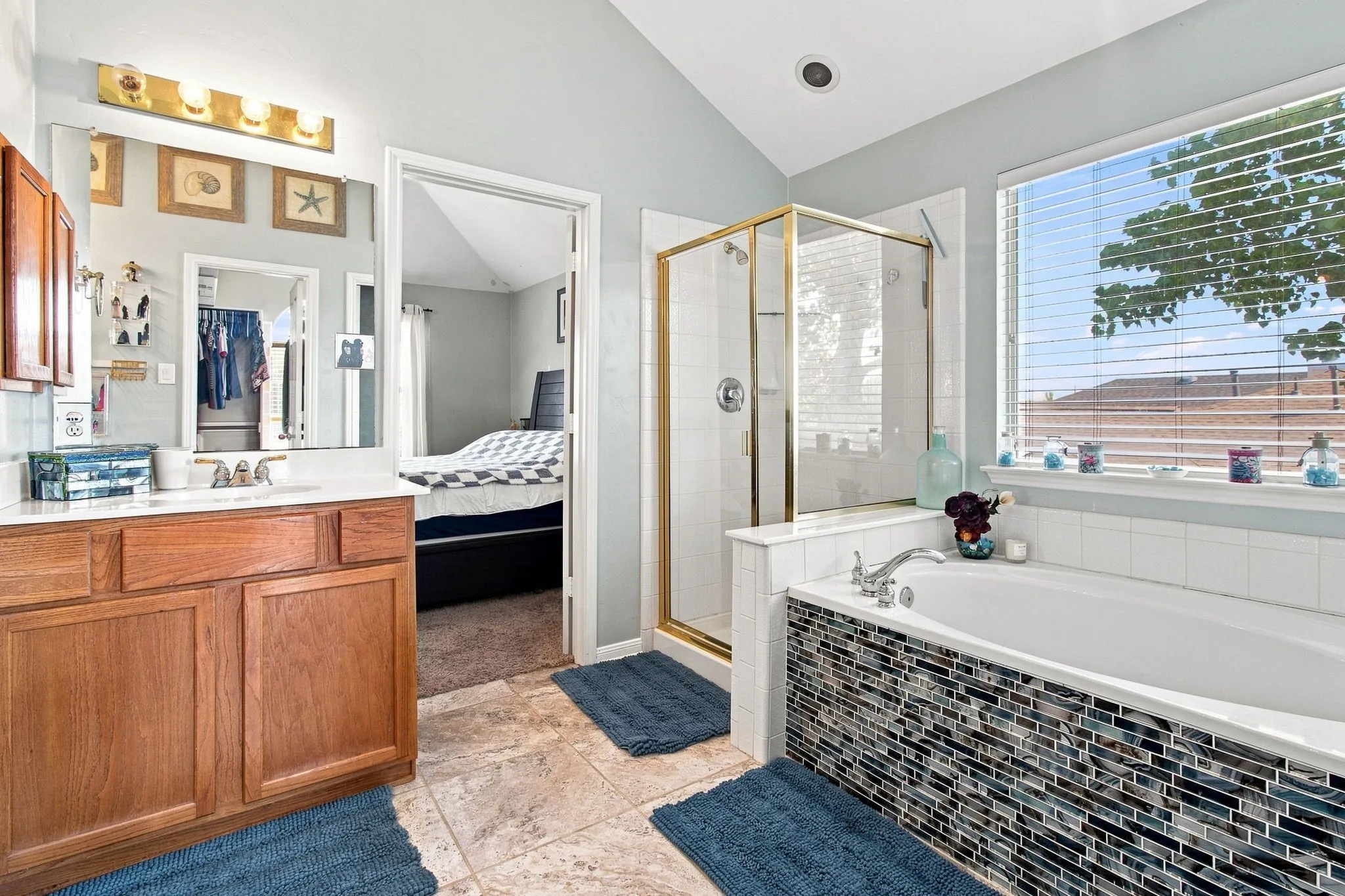 Dual vanity with integrated sink, glass-enclosed shower, soaking tub with mosaic tile surround, large window with blinds, and vaulted ceiling