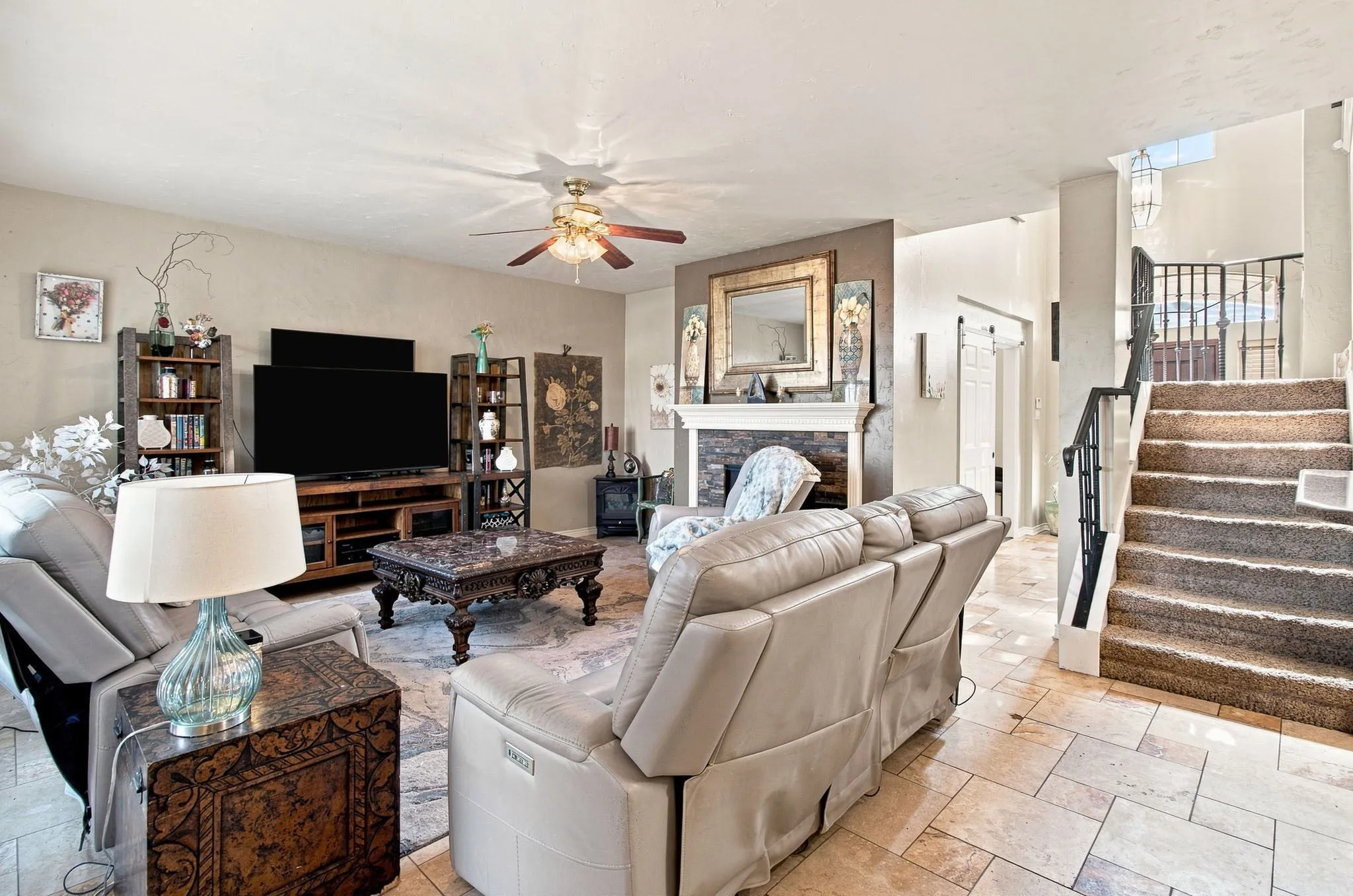 Living area featuring a stone-clad fireplace with a mantel, tile flooring, a ceiling fan, and a staircase with a wrought-iron railing