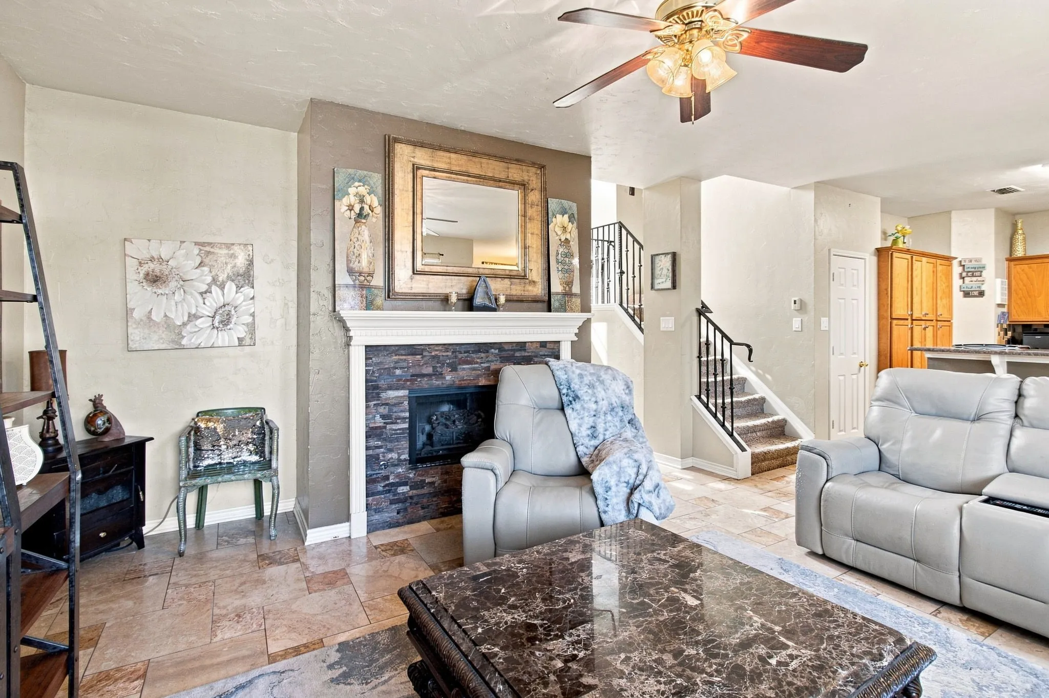 Stacked stone fireplace with a white mantel, tiled flooring, ceiling fan with light fixture, and a staircase with wrought iron balusters
