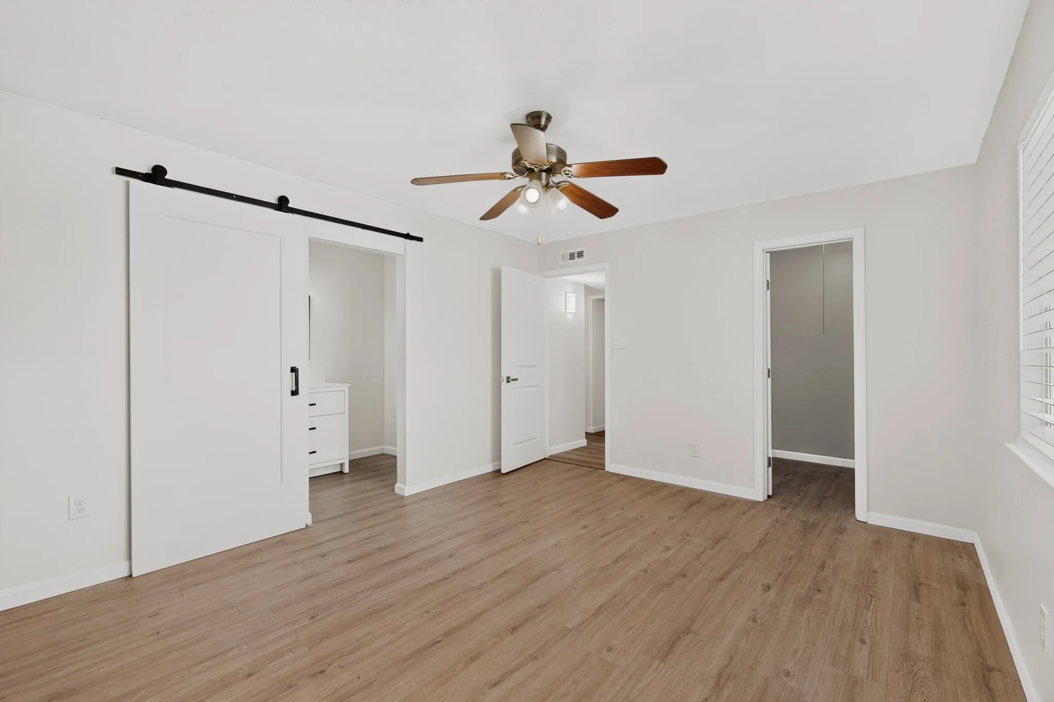 Wood-finish flooring, ceiling fan with light fixture, window with blinds, white walls, and a barn door with black hardware