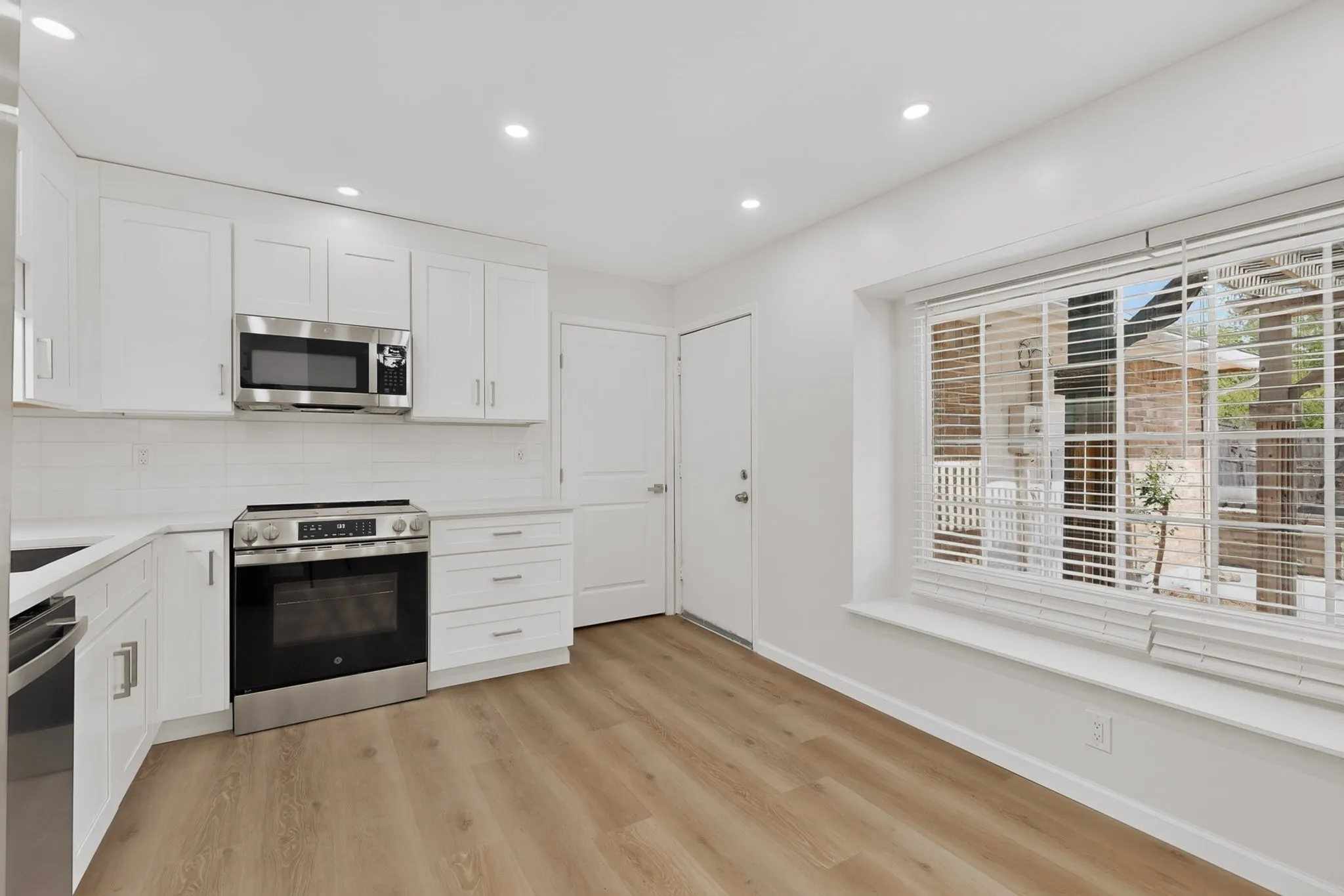 White cabinetry, stainless steel appliances, white subway tile backsplash, light wood-finish flooring, and a large window with blinds