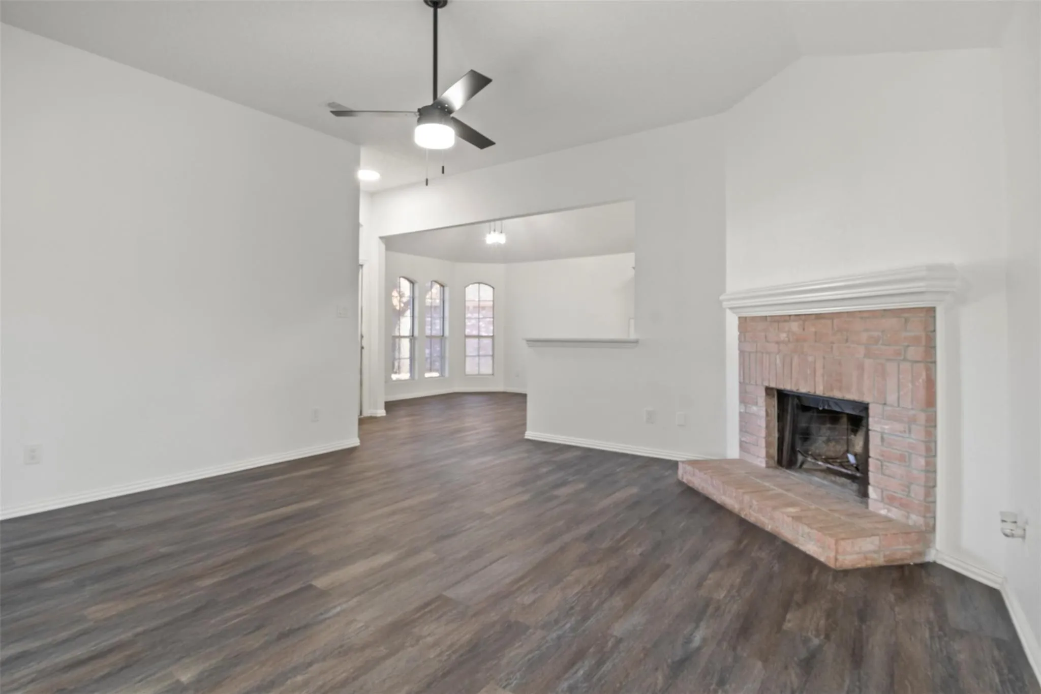 Wood-finish flooring throughout, brick fireplace with mantel, ceiling fan with integrated lighting, and a pass-through opening to an additional room with bay windows