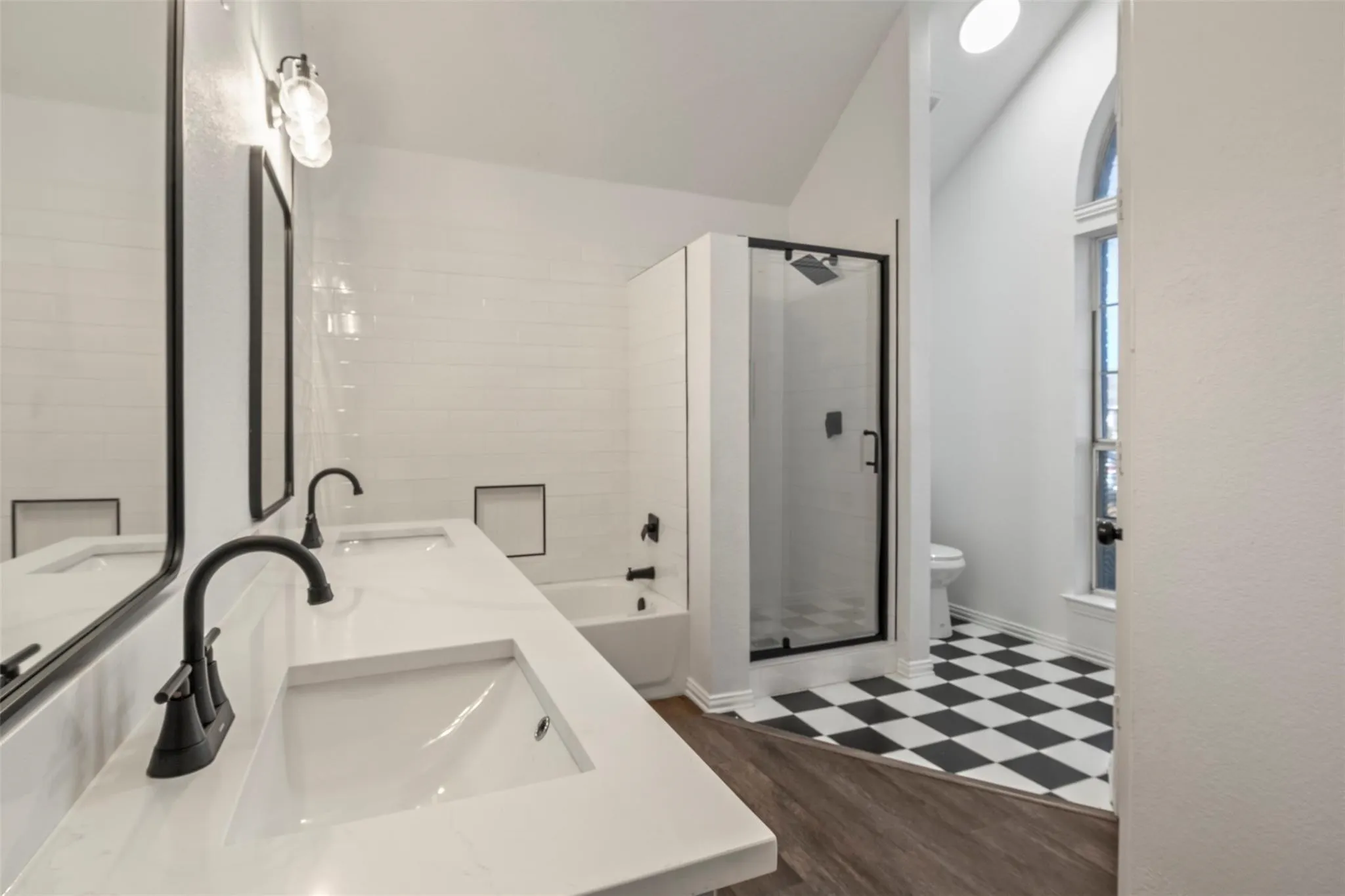 Dual vanity with integrated sinks and black matte fixtures, glass-enclosed shower with black trim, white subway tile surround, checkered floor tile, and arched window